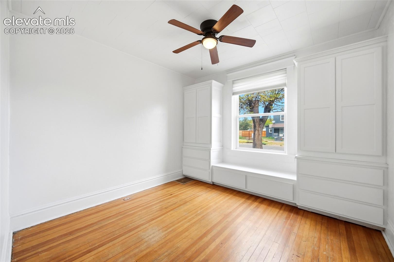 Wood-finish flooring, white built-in cabinetry featuring drawer storage, integrated window seat, and a ceiling fan with light fixture