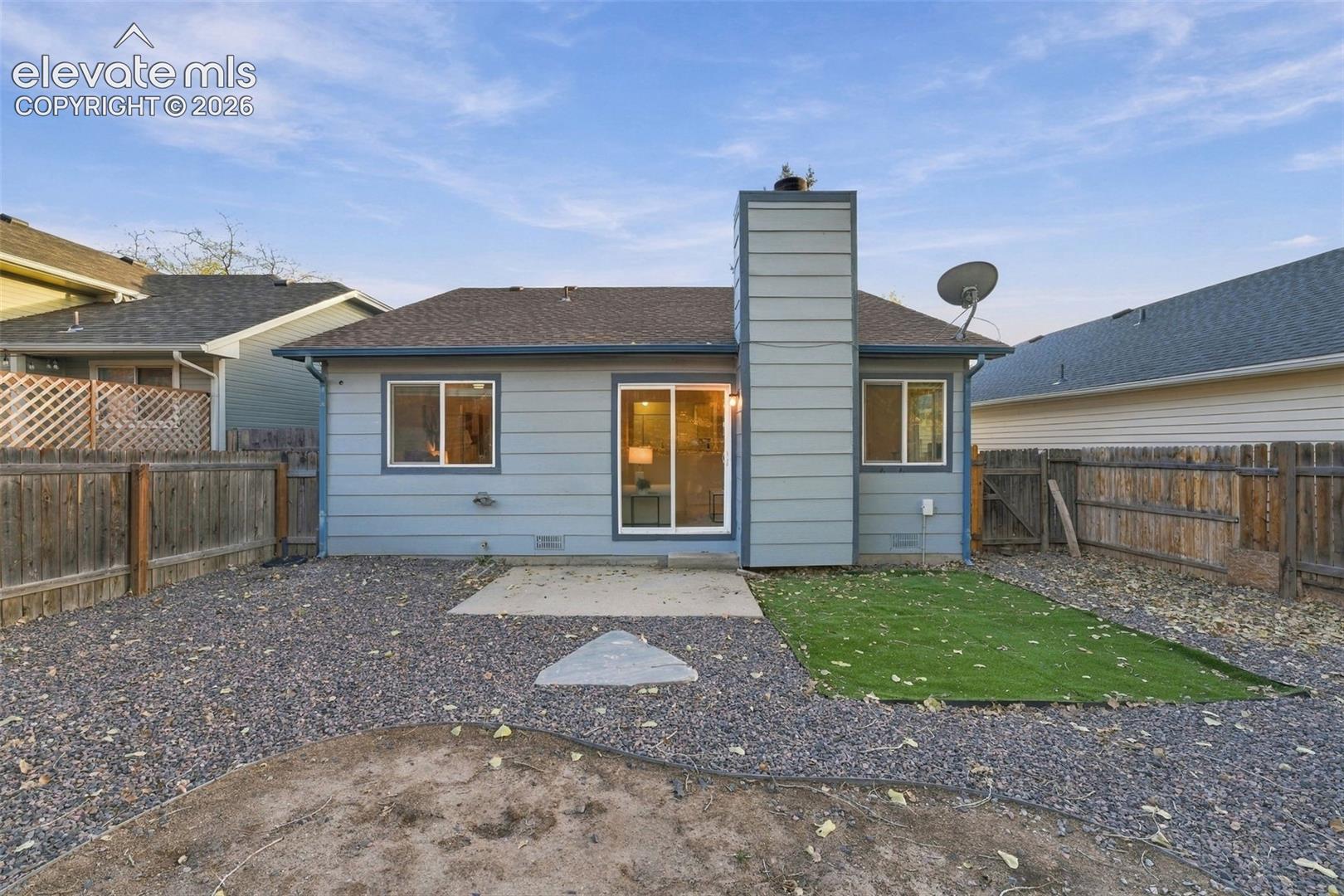 Rear exterior featuring horizontal lap siding, a prominent vertical chimney stack, a sliding glass door, a concrete patio, and a fenced-in yard with a gravel and artificial turf area