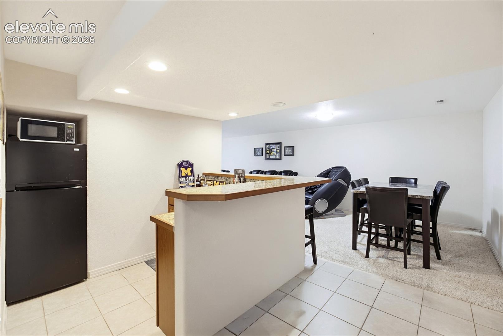 Recessed lighting, kitchen bar with wood-finish countertop, black refrigerator, built-in microwave nook, and neutral-toned tile flooring