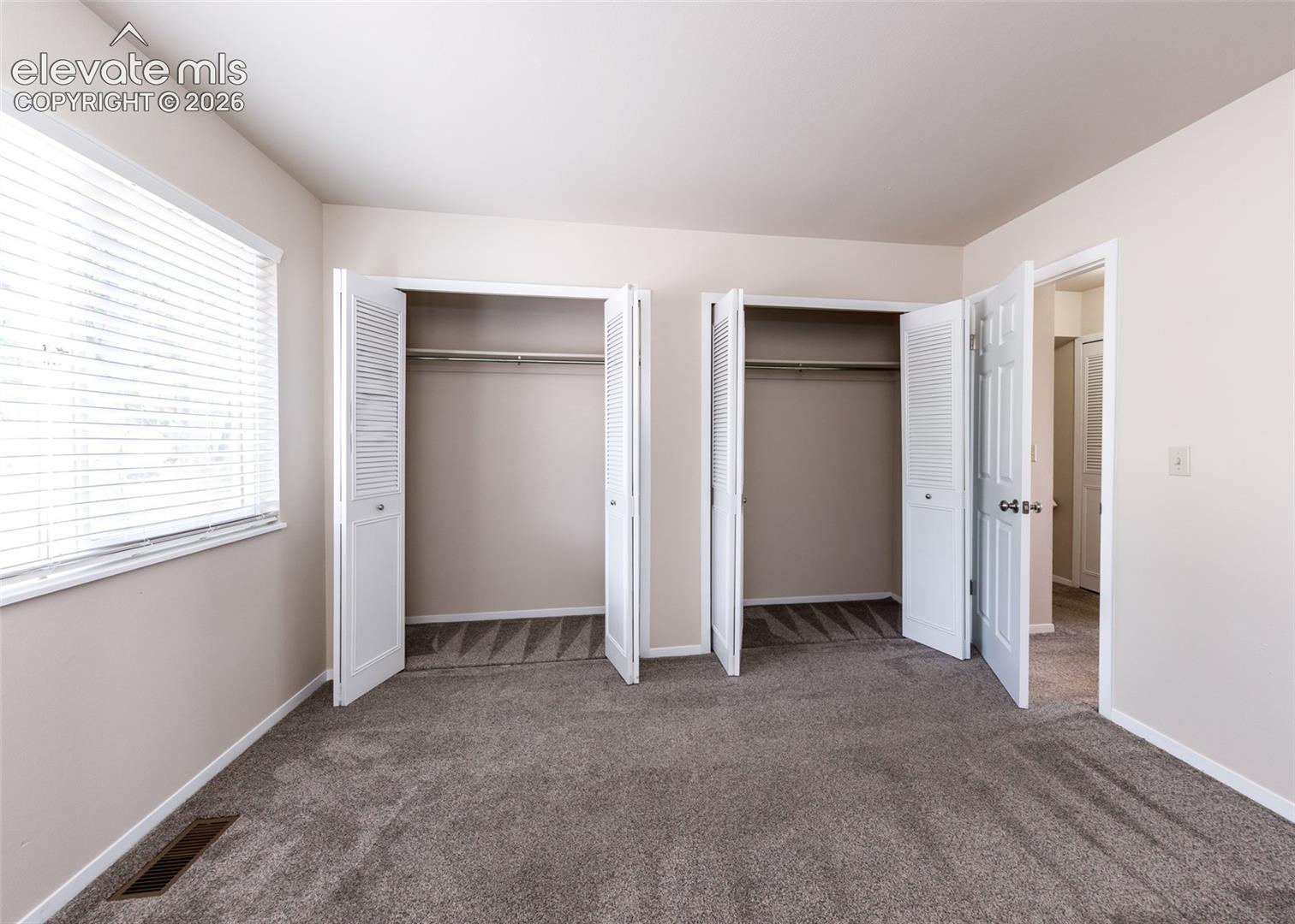 Plush gray carpeting, dual bi-fold closets, window with horizontal blinds, neutral wall paint