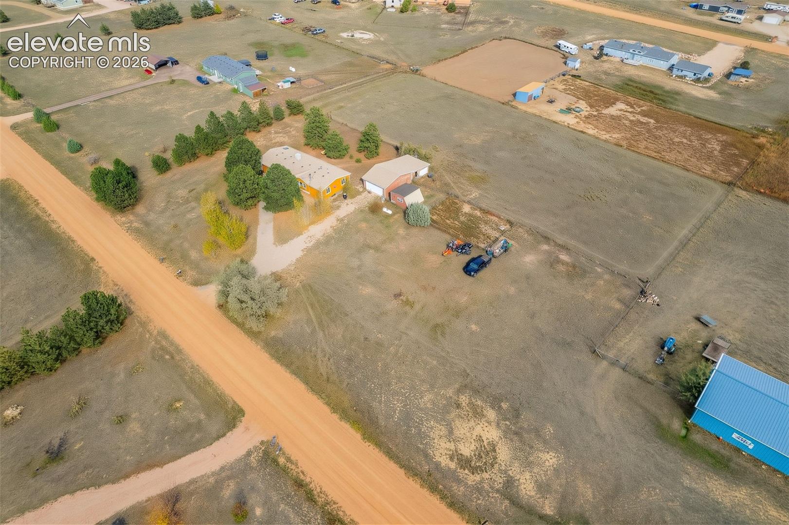 Aerial perspective of a multi-structure property featuring a detached garage, expansive acreage, mature tree clusters, and a paved driveway