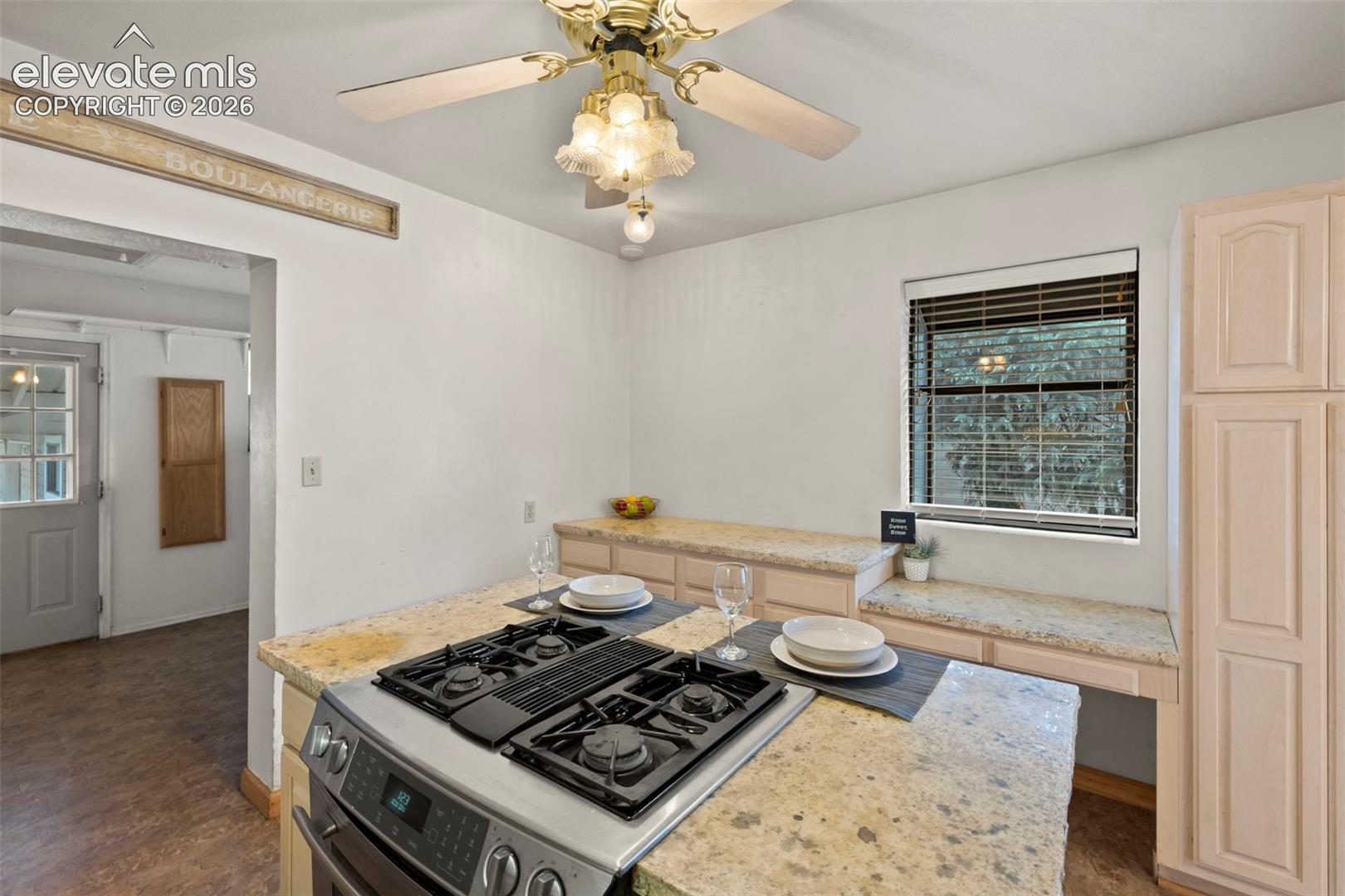 Granite countertops, built-in cooktop, light wood-finish cabinetry, ceiling fan with light fixture, and window with blinds