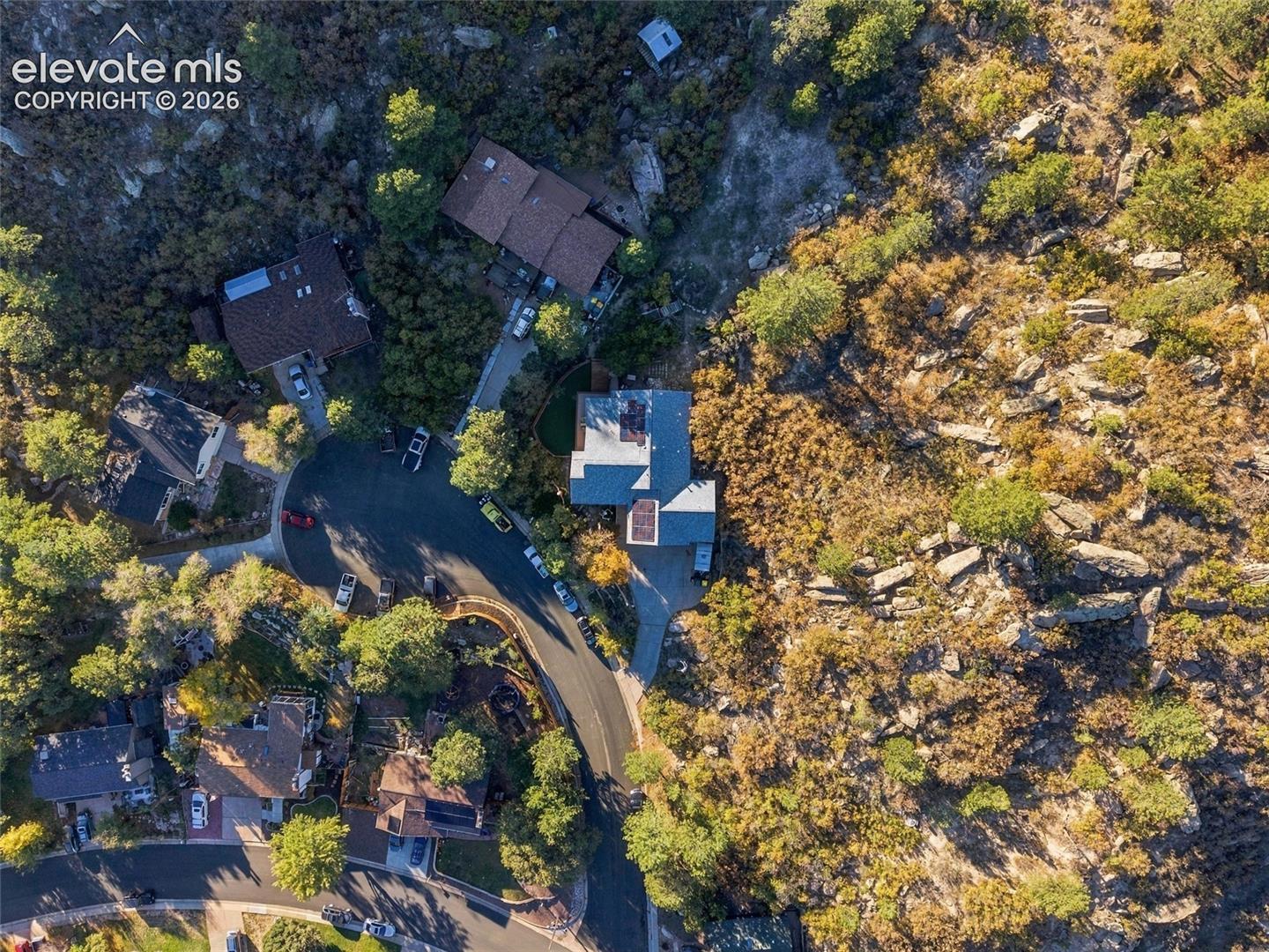 Aerial perspective featuring a multi-level structure, paved driveway, solar panel installation, and expansive natural rock landscape with mature evergreen trees