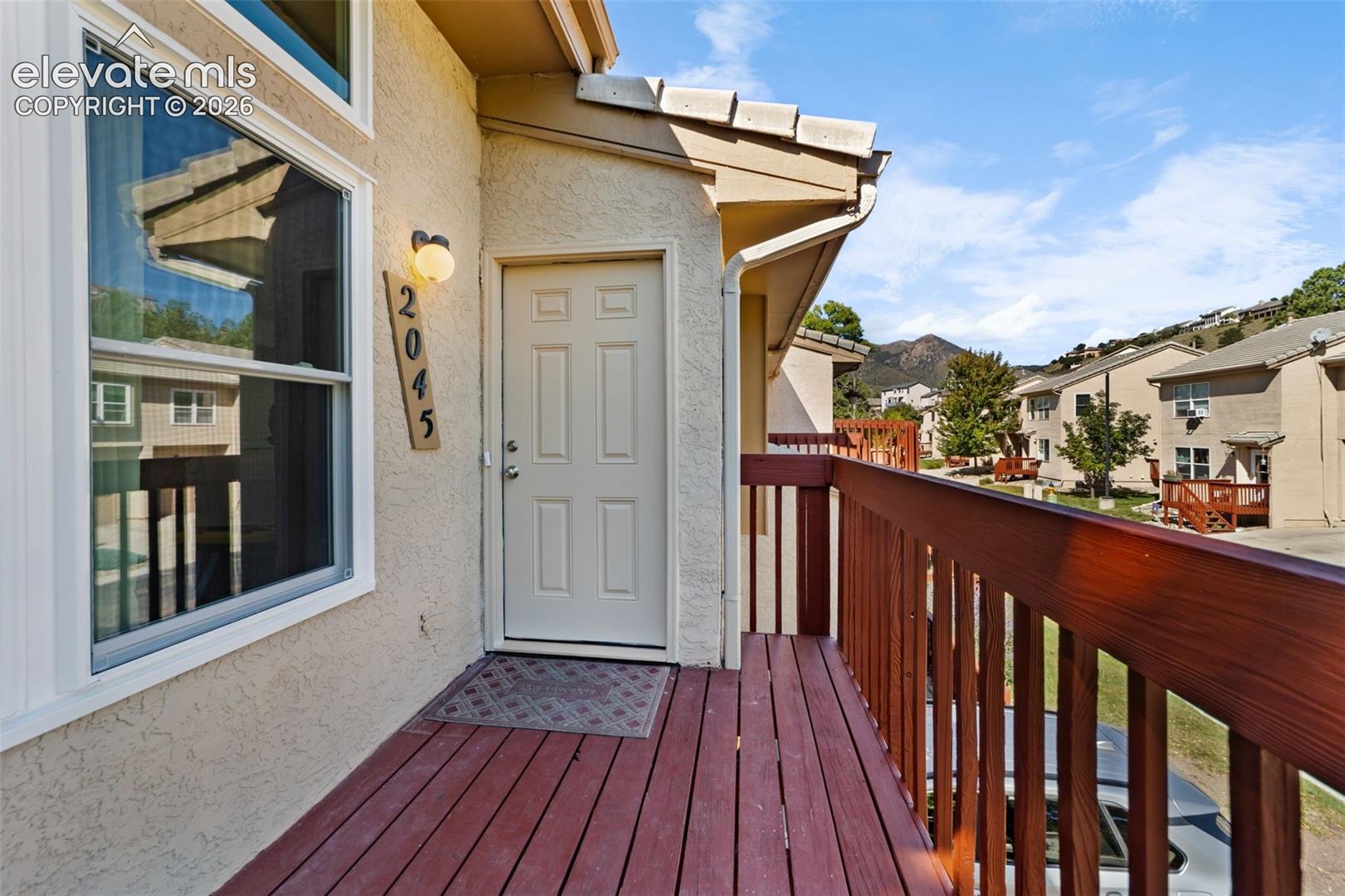 Private balcony with wooden decking and balustrade, exterior light fixture, and six-panel entry door featuring a mountain backdrop