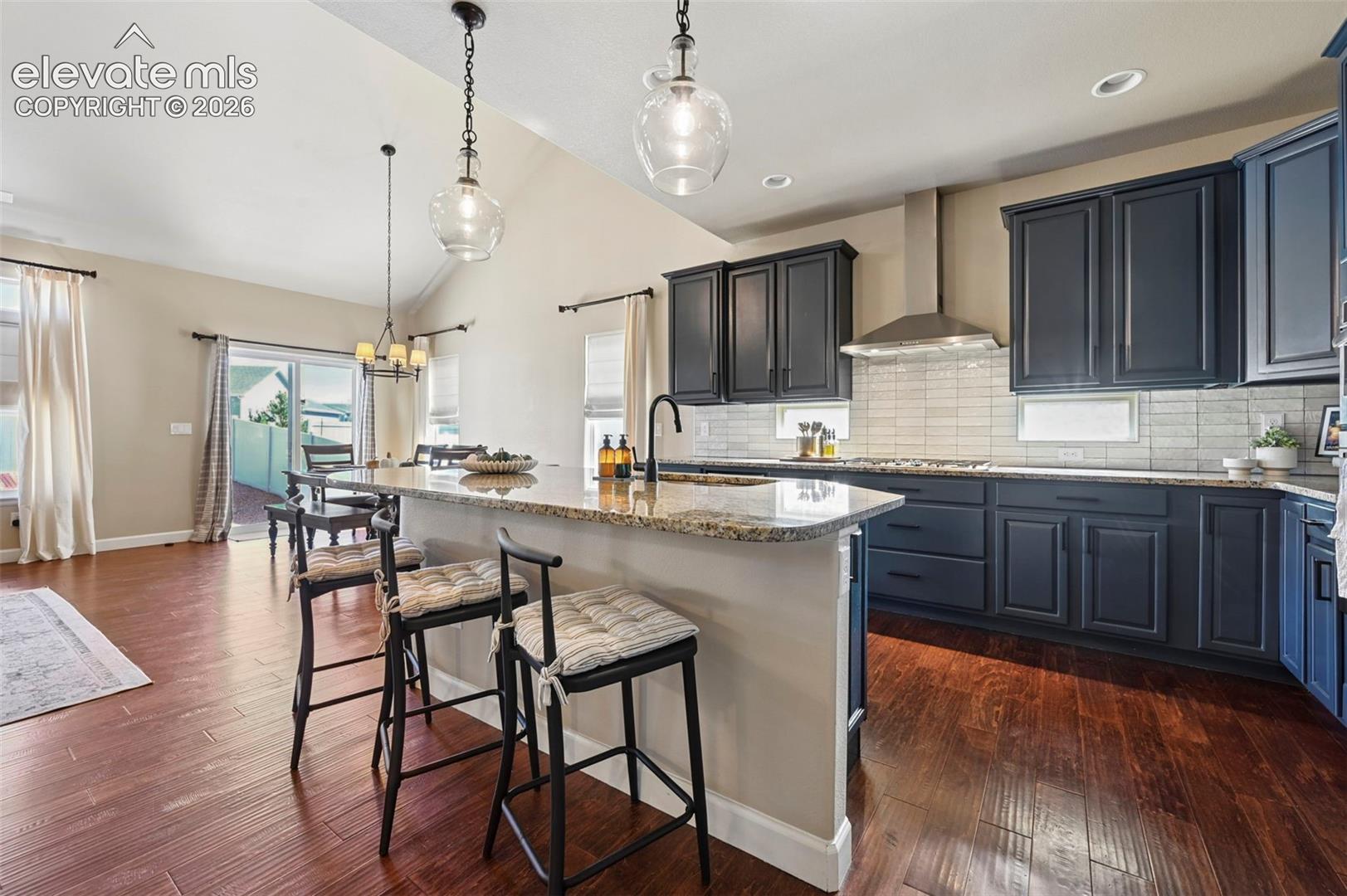 Kitchen featuring wood-finish flooring, dark-toned cabinetry, stone-surfaced island, subway tile backsplash, and stainless steel range hood