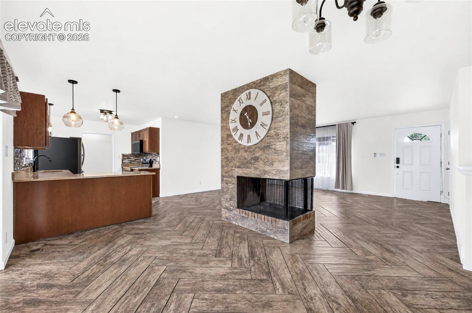 Herringbone wood-finish flooring, central dual-sided stone-surround fireplace, kitchen peninsula with cabinetry, and multi-light hanging fixtures