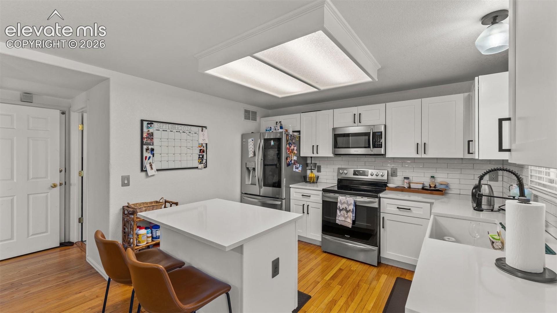 Kitchen featuring wood-finish flooring, white cabinetry, integrated stainless steel appliances, white subway tile backsplash, and a white island with countertop seating