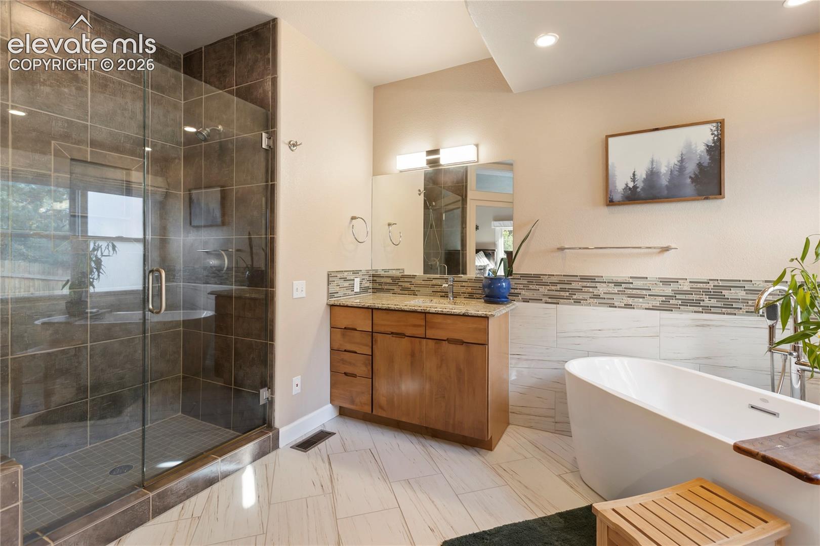 Glass-enclosed shower with dark tile surround, freestanding soaking tub, wood-finish vanity with countertop, and mosaic tile backsplash