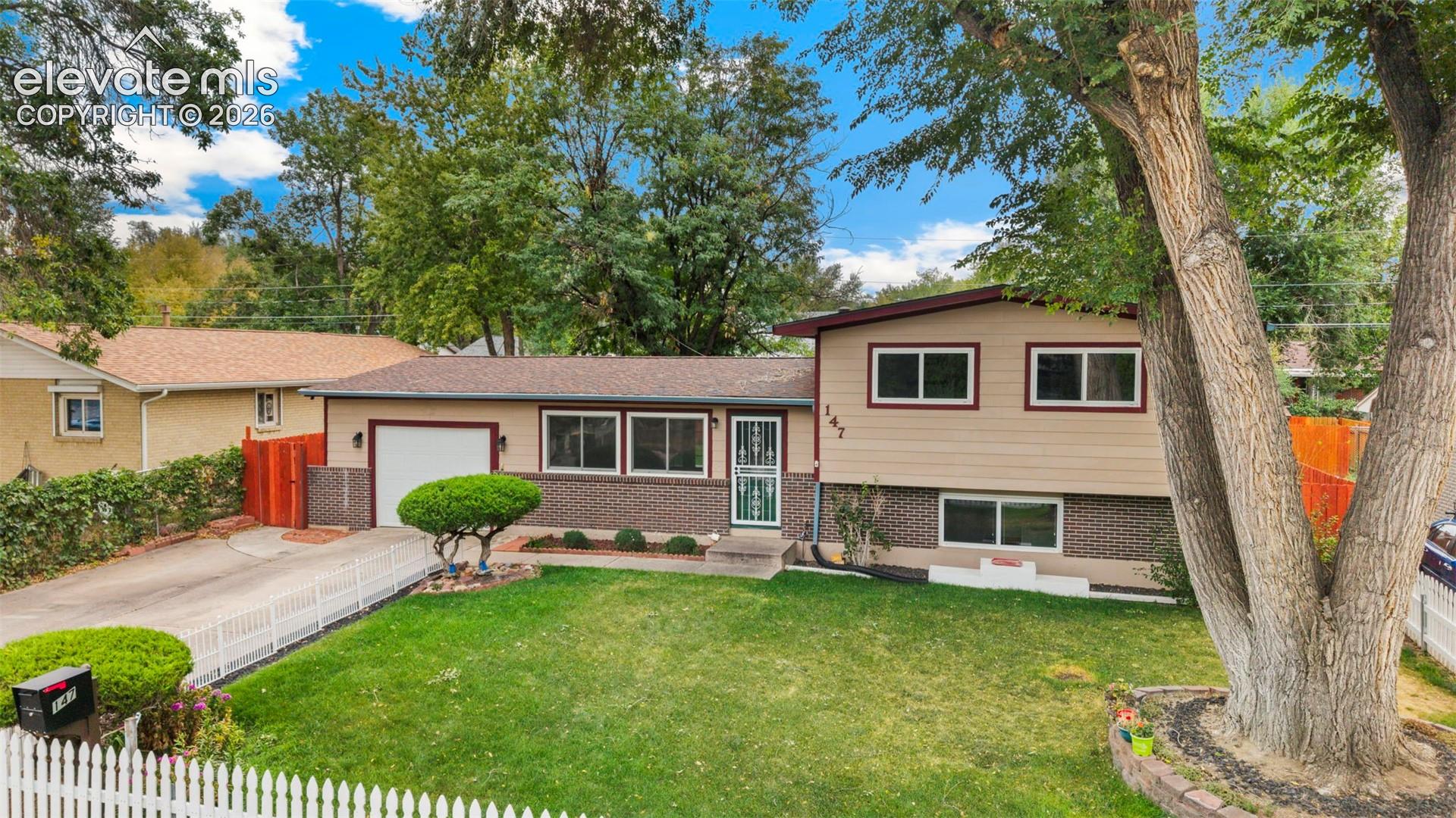 Split-level residence featuring a two-tone facade with brick detailing, an attached garage, a white picket fence, and a manicured lawn