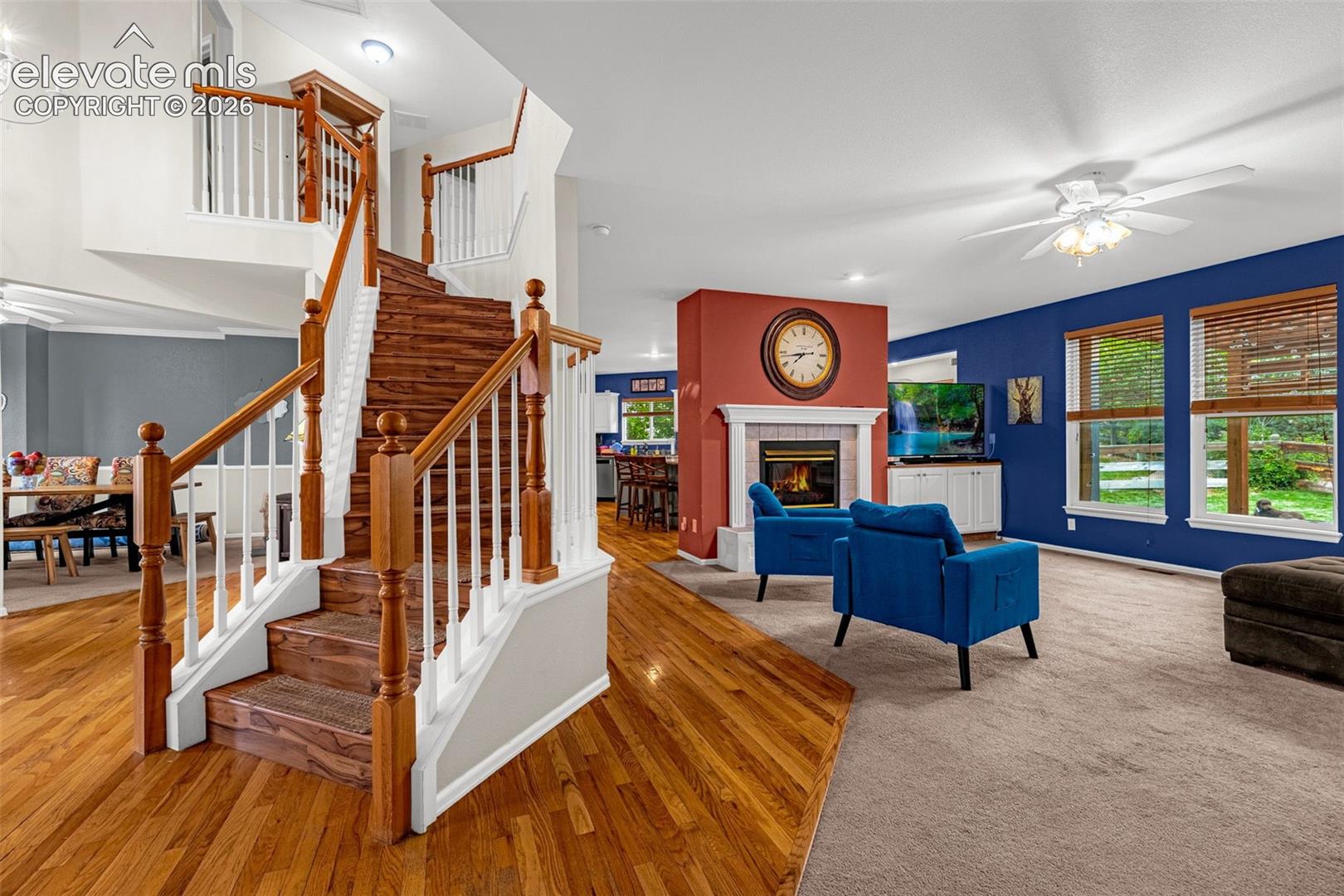 Hardwood flooring, two-story staircase with wood newel posts, built-in fireplace with mantel, multiple large windows, and a ceiling fan
