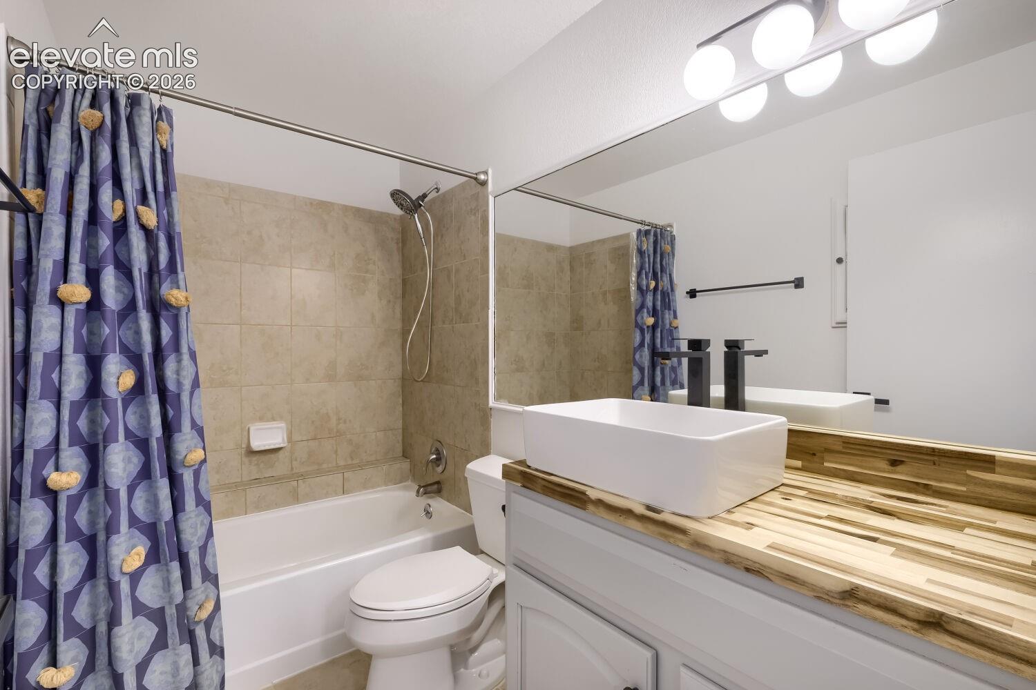 Bathroom featuring a white rectangular vessel sink, wood-finish countertop, matte black faucet, wall-spanning vanity mirror, and a tiled shower-tub surround