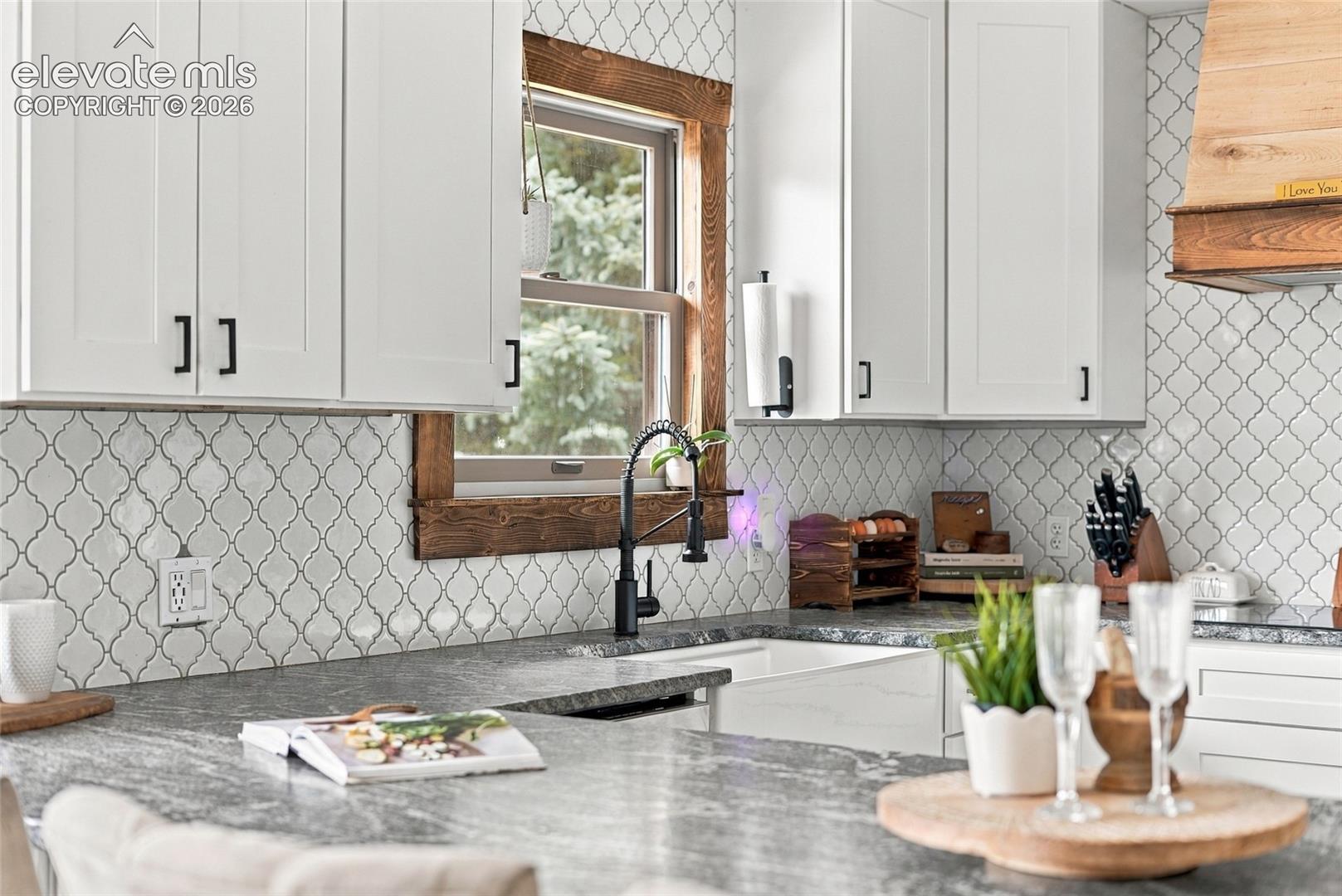 White shaker-style cabinetry with black hardware, Arabesque tile backsplash, wood-framed window, black gooseneck faucet, and integrated sink