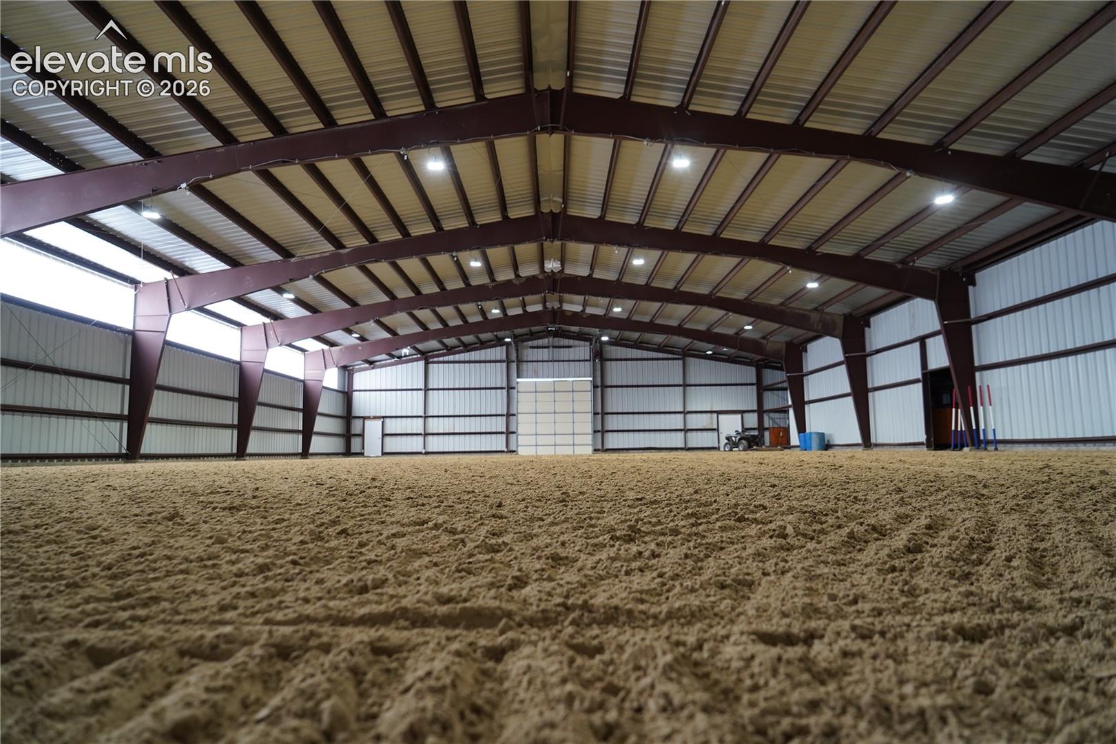Expansive indoor arena with a sand footing, high ceilings with exposed steel beams, and multiple overhead light fixtures