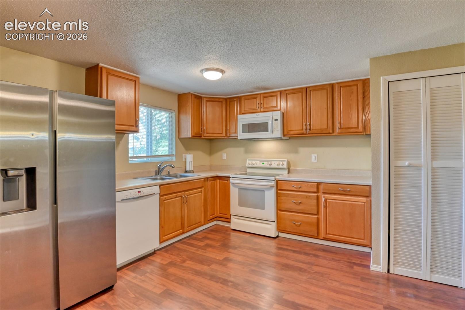 Kitchen featuring wood-finish flooring, a full suite of appliances including a stainless steel refrigerator, white dishwasher, and white range with microwave above, light-toned countertops, and ample wood cabinetry