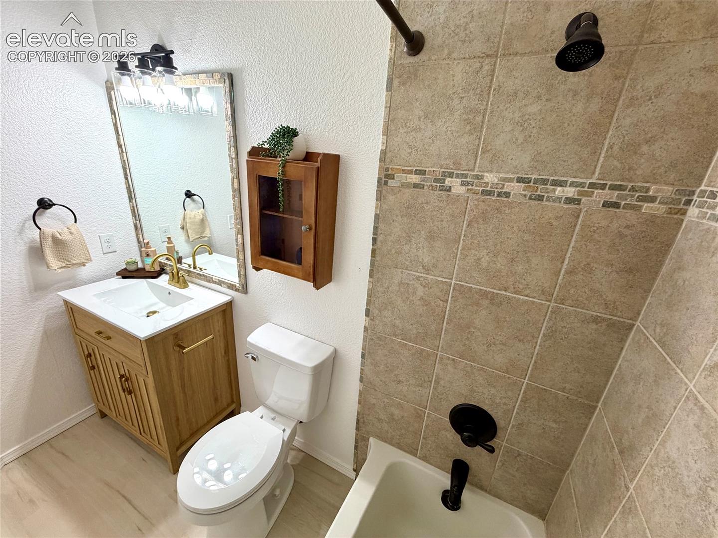 Upstairs guest bathroom featuring a light wood-finish vanity with a white countertop and gold-finish faucet, a framed mirror, and a ceramic tile shower surround with a decorative mosaic accent band