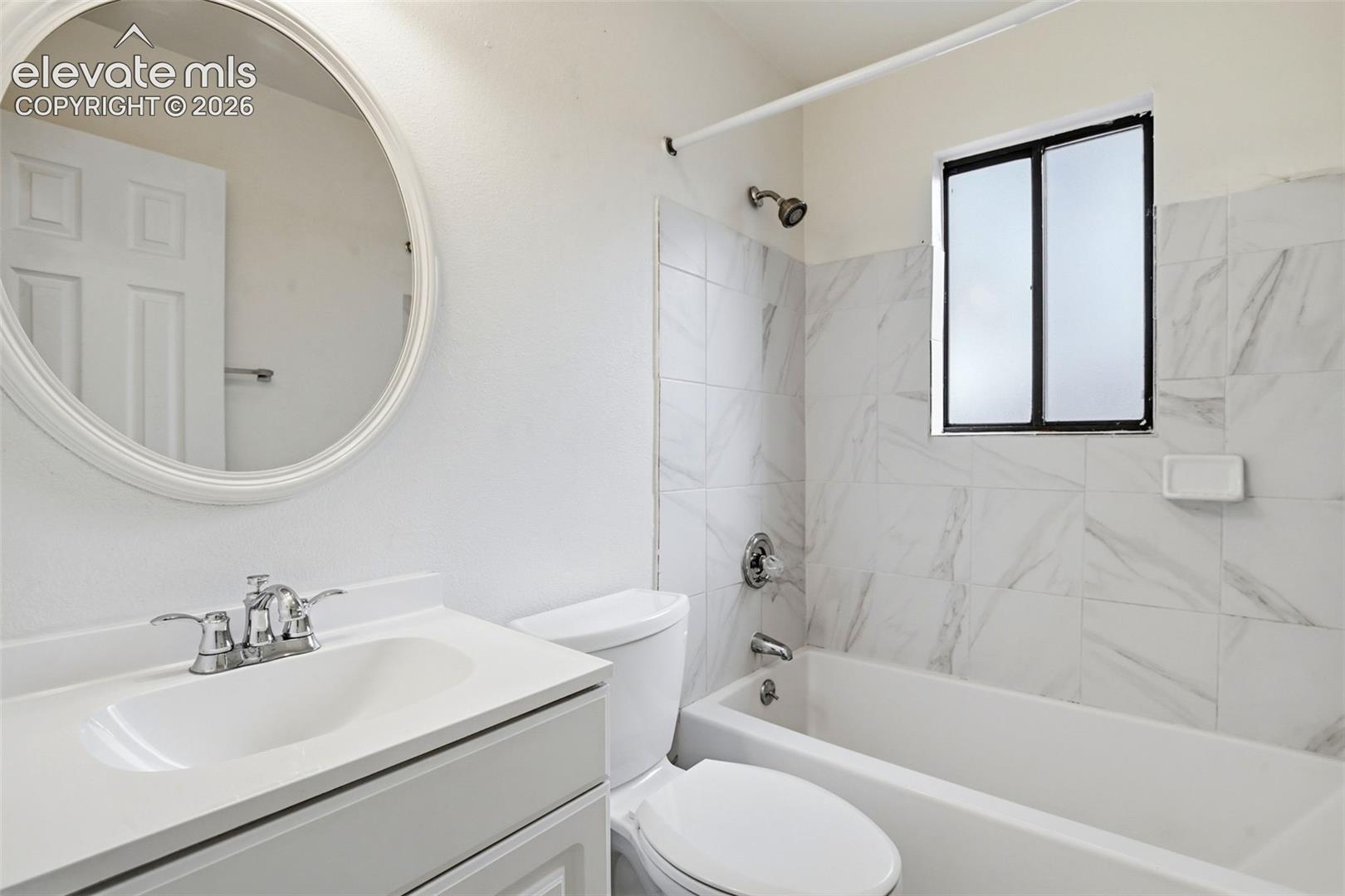 Bathroom featuring a white vanity with integrated sink, chrome faucet, oval wall mirror, full-size bathtub with shower, and white tile surround