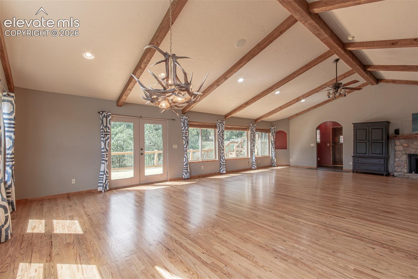Vaulted ceilings with exposed wood beams, wood-finish flooring, floor-to-ceiling windows, antler-style chandelier, and stone-surround fireplace