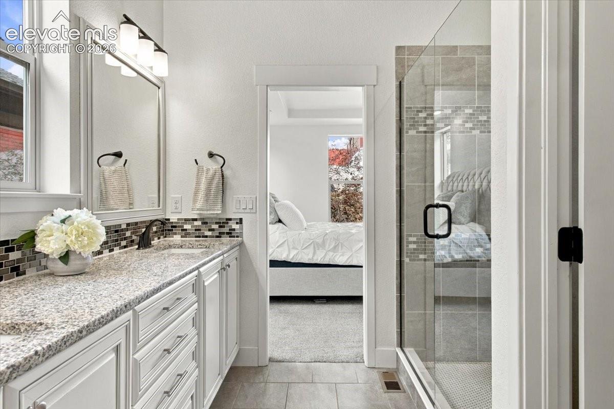 Bathroom featuring a dual-sink vanity with stone-finish countertops, mosaic tile backsplash, glass-enclosed shower with tile surround, and framed vanity mirror with overhead lighting