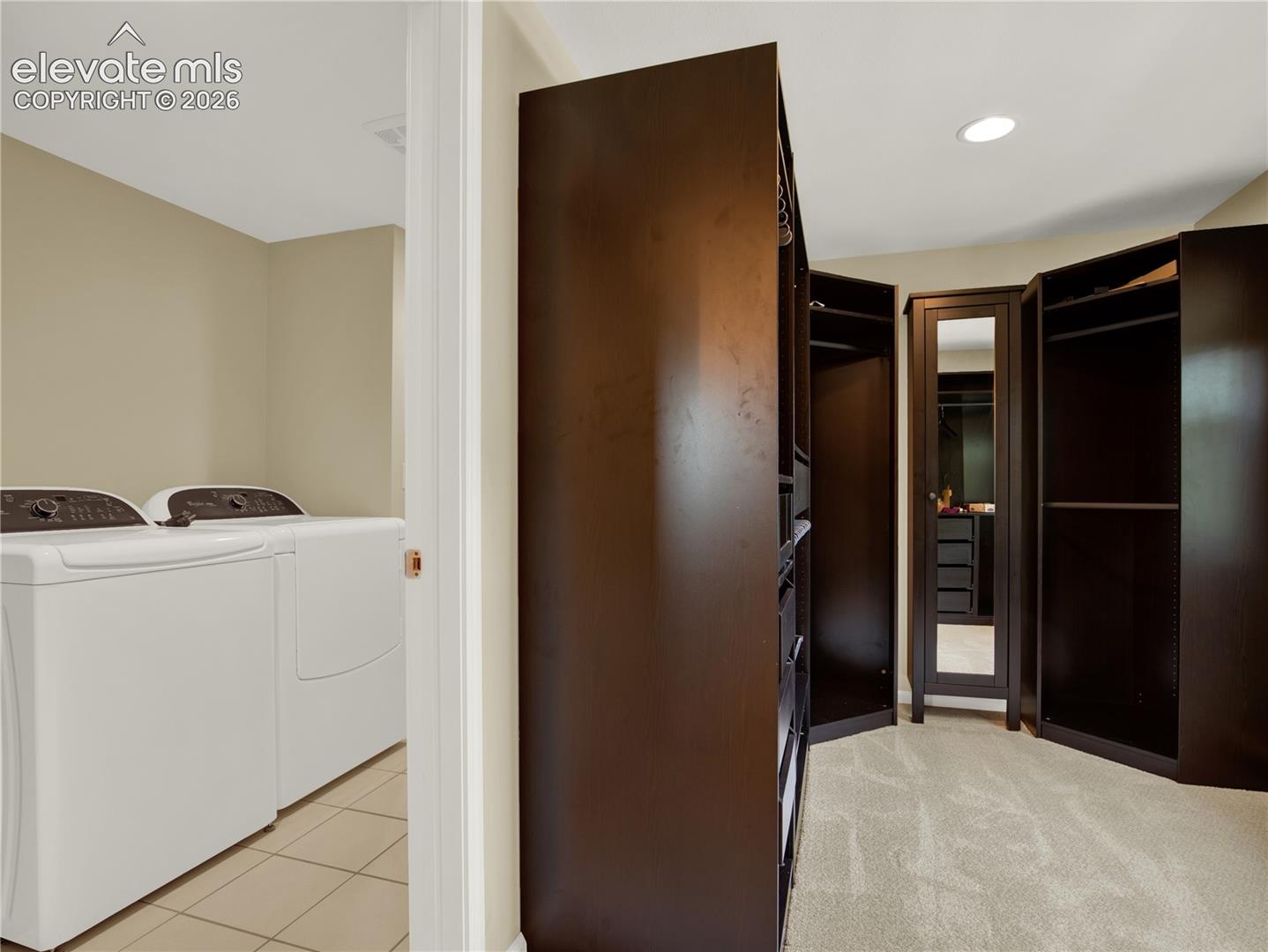 Walk-in closet featuring dark wood-finish built-in cabinetry, integrated mirrored panel, and recessed lighting, paired with a laundry area including tiled flooring