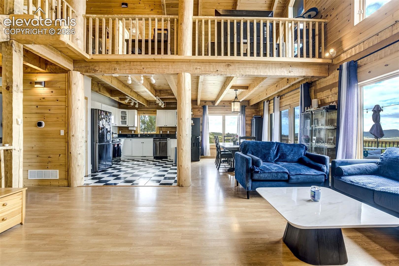 Expansive open-concept living space featuring wood-finish flooring, exposed timber beams, a full kitchen with checkered flooring and white cabinetry, a dedicated dining area, and a second-story loft with log railings