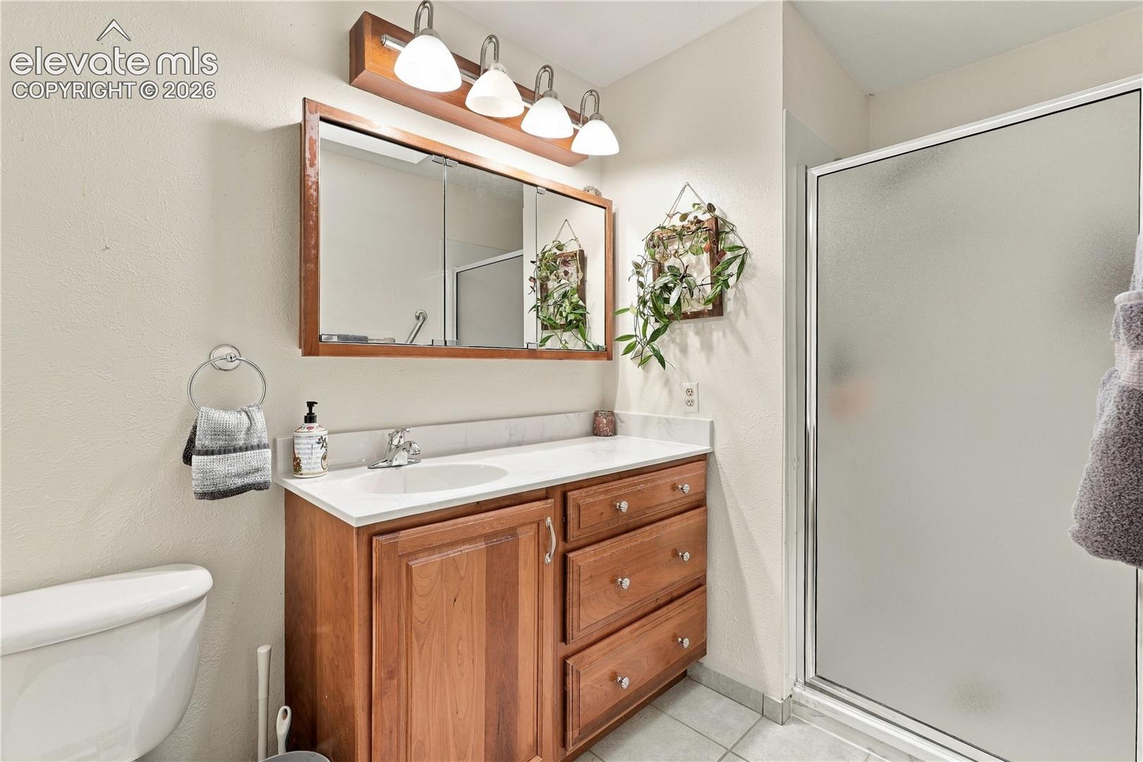 Wood-finish vanity cabinet, solid-surface countertop with integrated basin, polished metal hardware, four-light wall fixture, and glass-enclosed shower