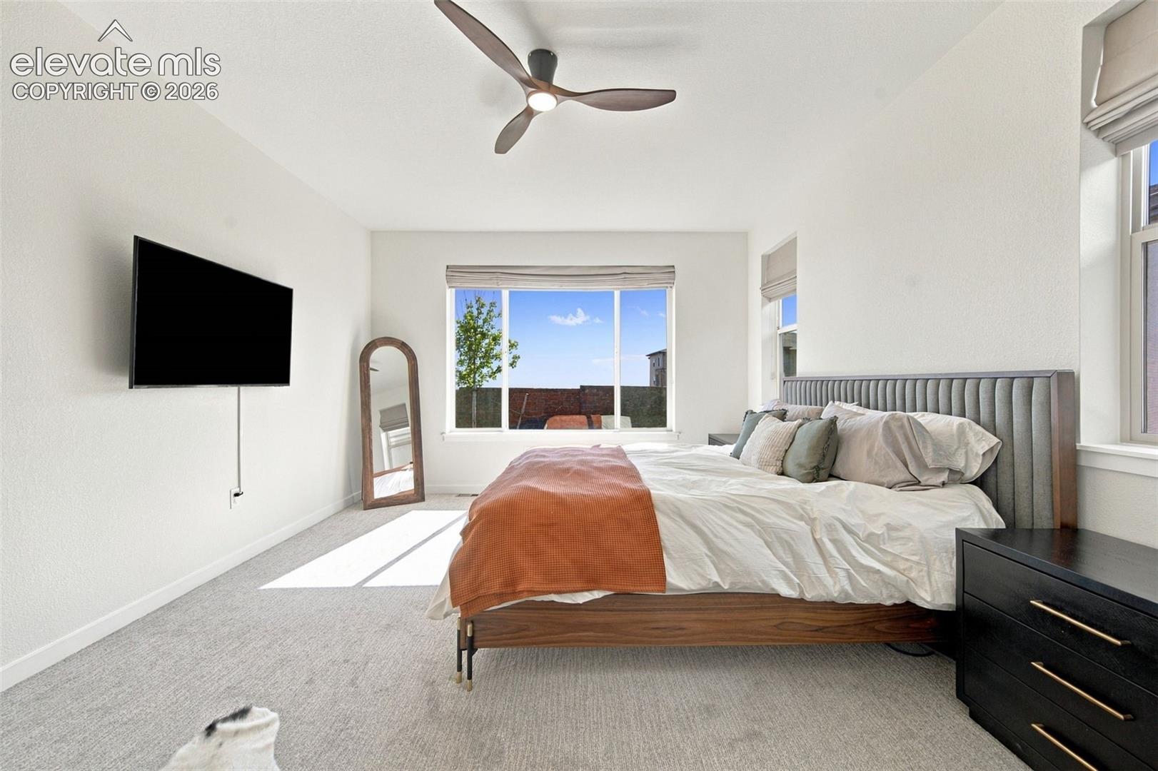 Neutral-toned carpeting, large picture window with light-filtering shade, and modern three-blade ceiling fan with integrated light