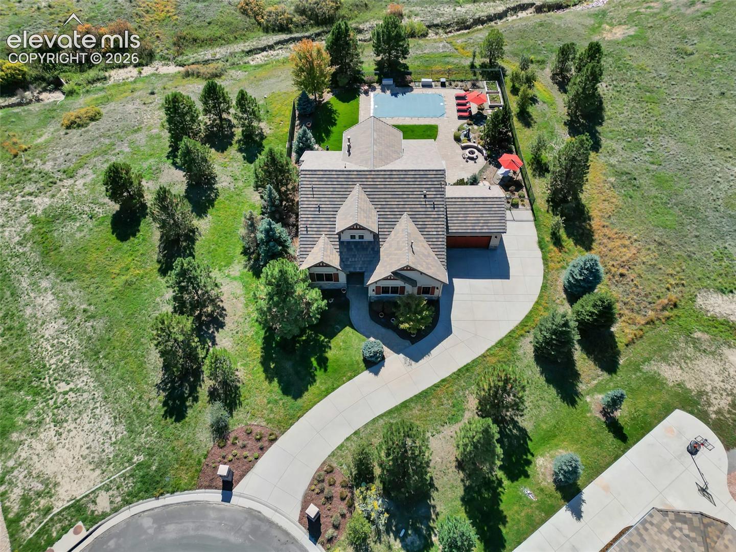 Aerial view of residential property featuring a multi-gabled shingle roof, paved driveway, in-ground swimming pool, stone-paver patio, and fire pit