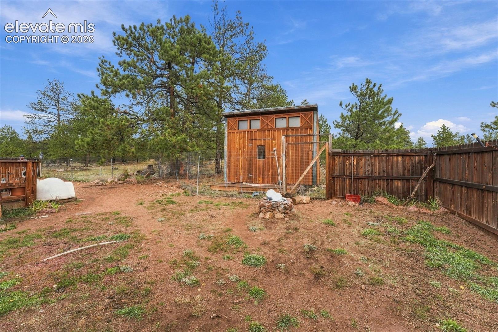 Detached wood-sided storage structure featuring a fenced yard, natural landscaping, and mature pine trees