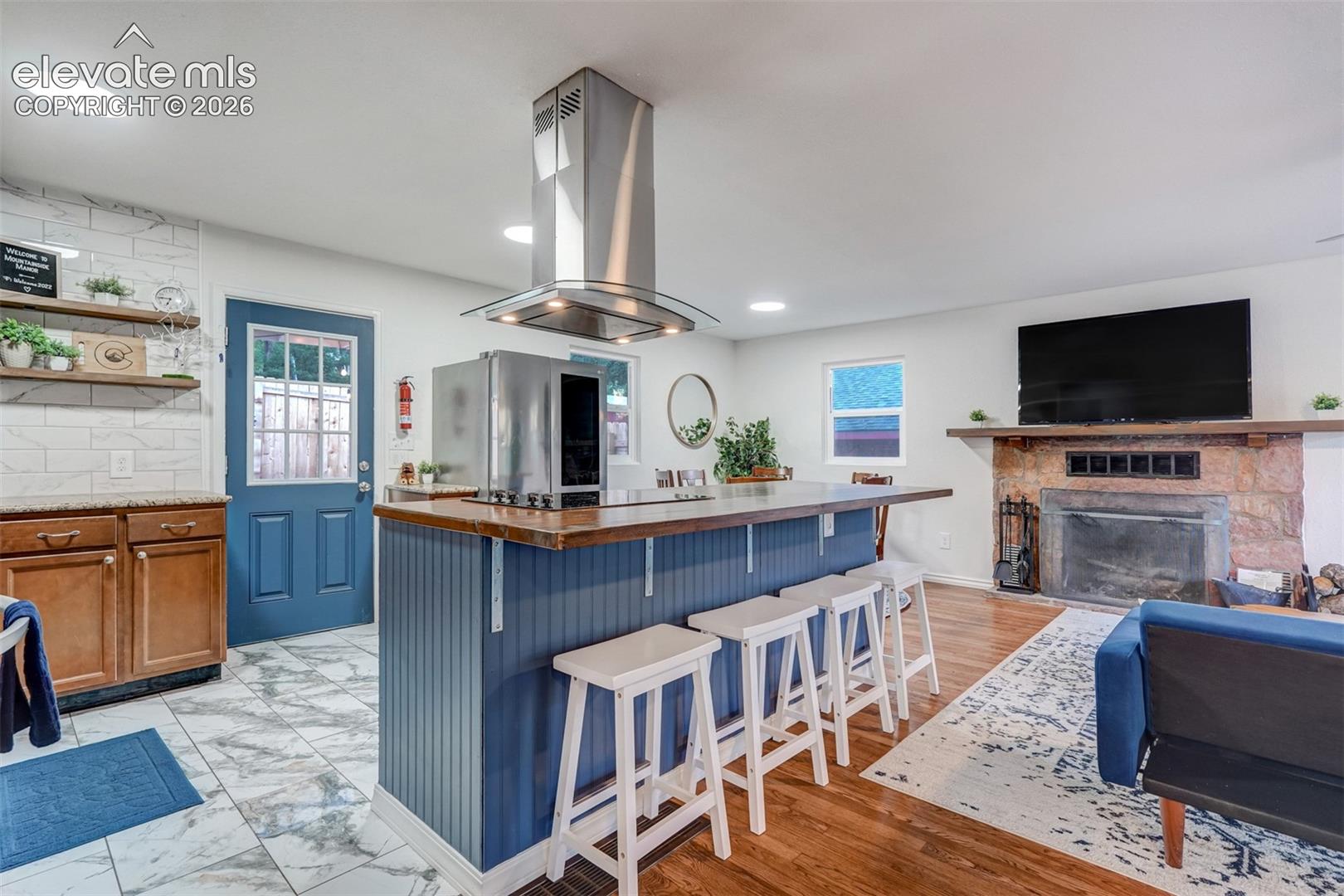 Open-concept living area featuring a kitchen island with seating, a stone fireplace, wood-finish flooring, and a blue paneled door