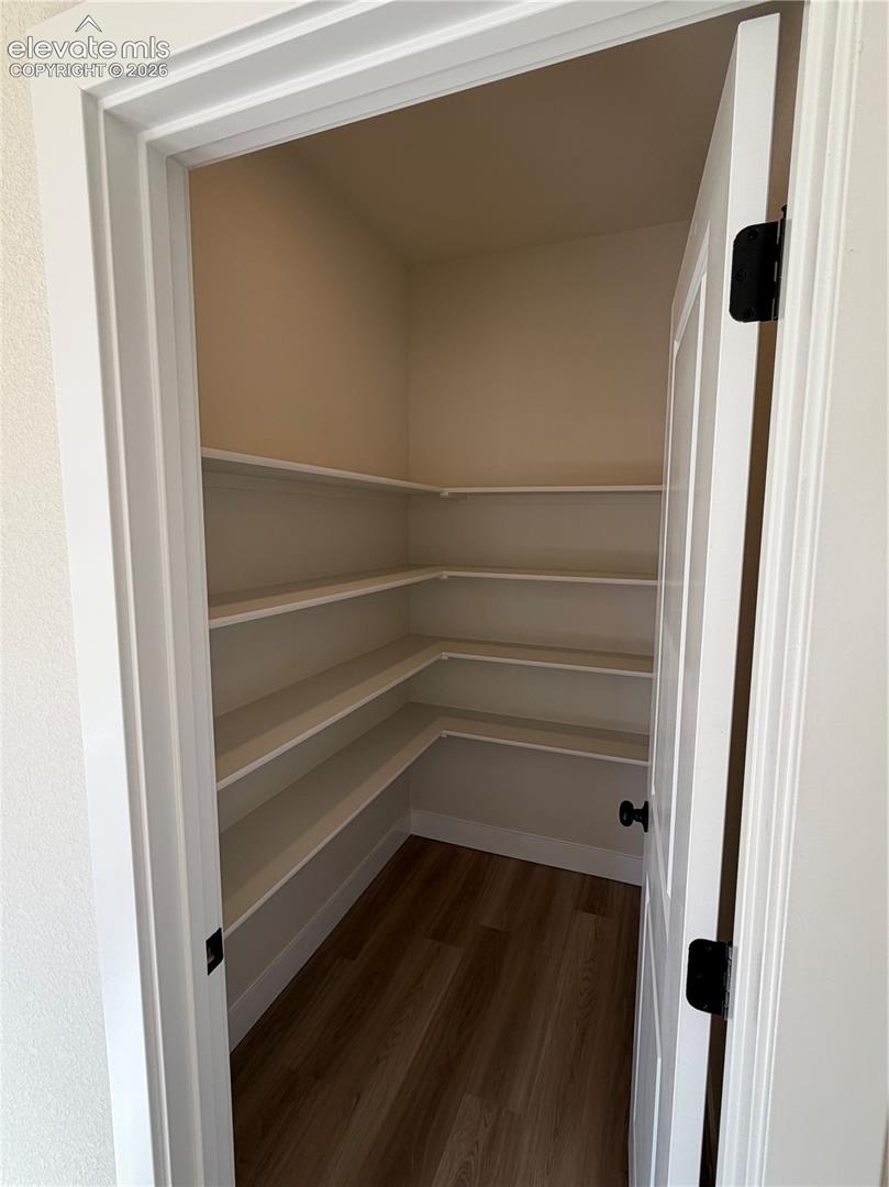 Walk-in storage space featuring multi-tiered white shelving, wood-finish flooring, and a door with matte black hardware