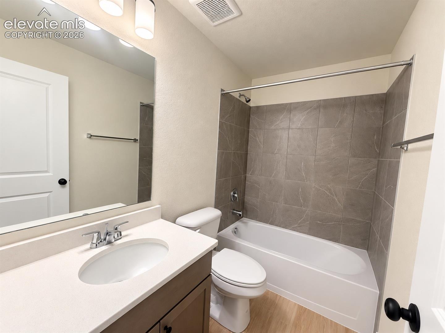 Vanity with a solid surface countertop, undermount oval sink, large wall-mounted mirror, soaking tub with tile surround, and wood-finish flooring