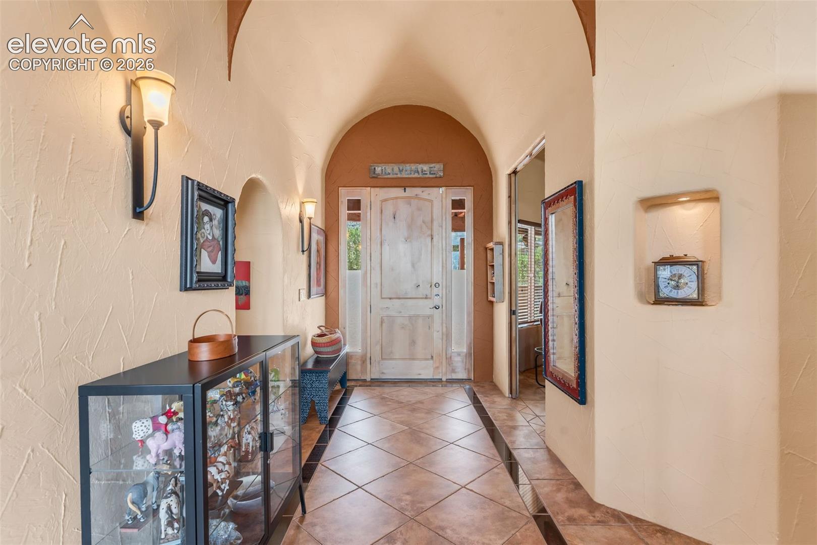 Entryway featuring a barrel-vaulted ceiling, diamond-patterned tile flooring with dark inlay borders, a solid wood entry door with side lites, wall-mounted light fixtures, and a recessed architectural wall niche