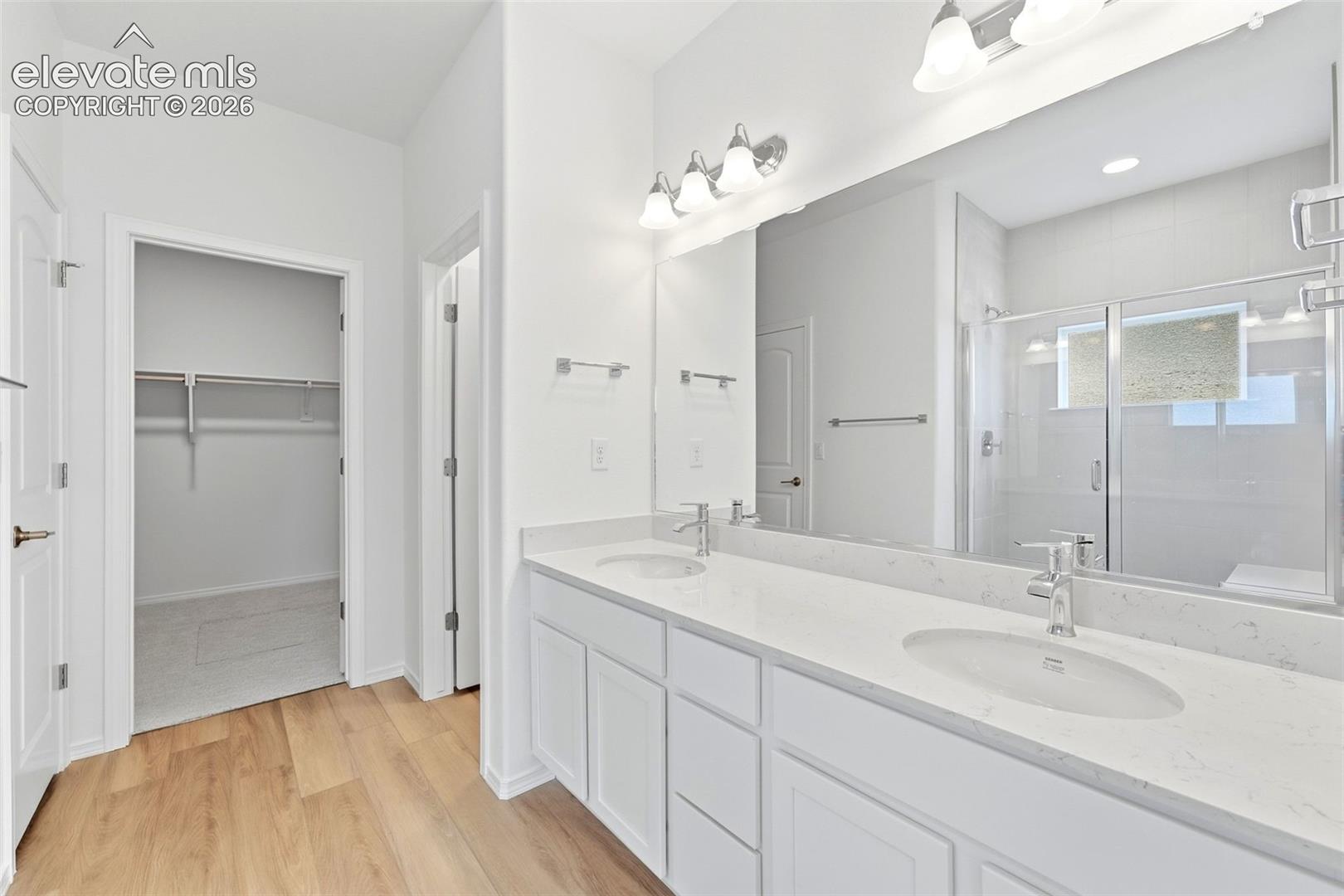 Bathroom featuring a dual-sink vanity with a solid-surface countertop, full-width mirror, wood-finish flooring, and a walk-in shower with a glass enclosure