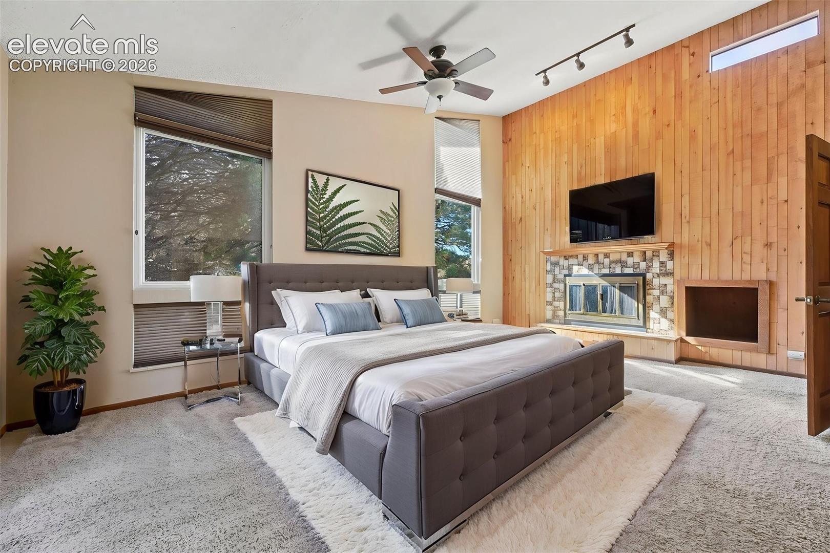 Vaulted ceilings, wood-paneled accent wall, fireplace with stone surround, ceiling fan, and multi-pane transom windows