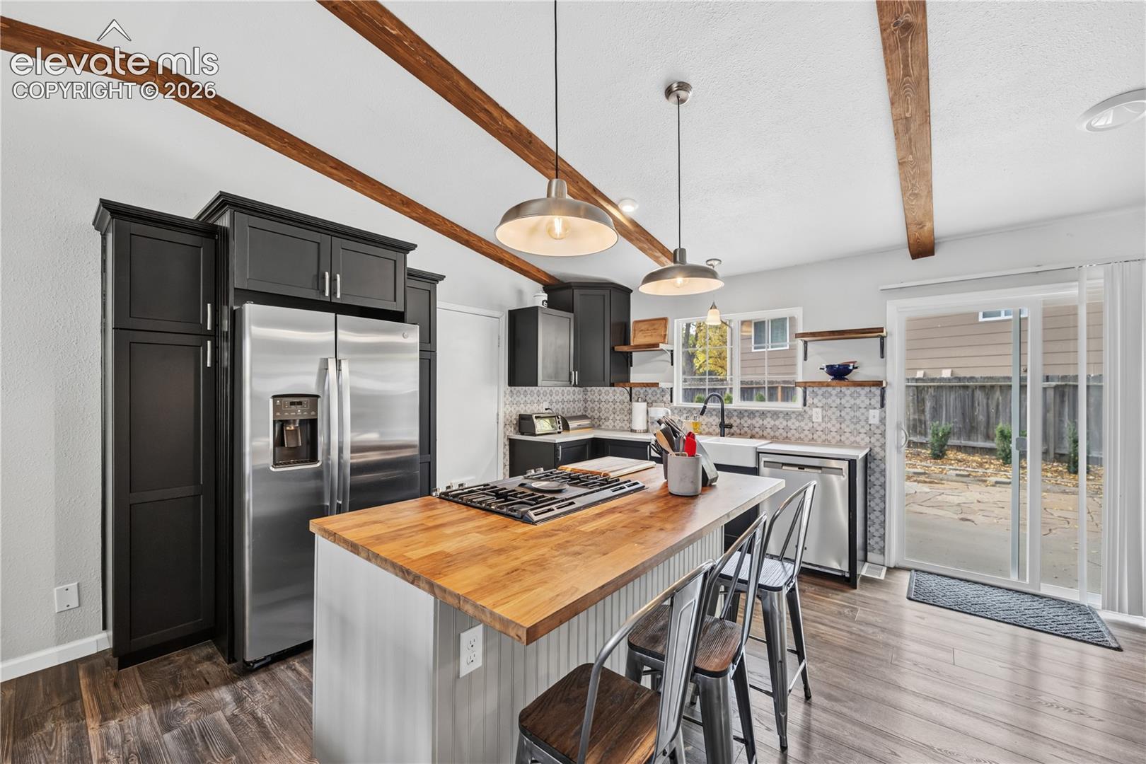Kitchen featuring a wood-topped center island with integrated gas cooktop, dark cabinetry, exposed ceiling beams, patterned backsplash, and an apron-front sink