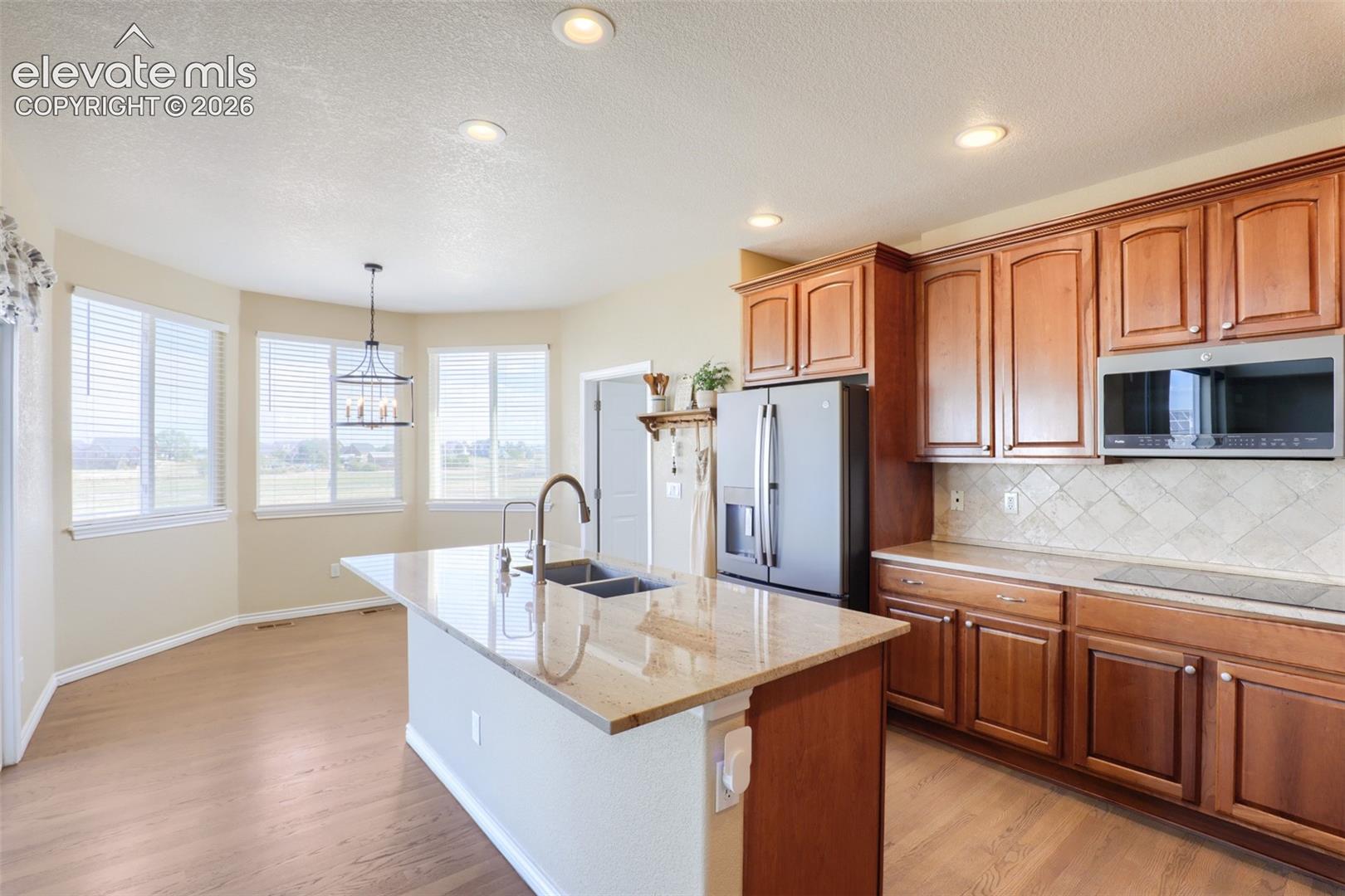 Well-appointed kitchen showcasing a sleek induction glass cooktop, granite countertops with a stone tile backsplash in a diagonal pattern, and stainless steel GE appliances, including a microwave hood and french door refrigerator. Rich wood cabinetry with crown molding frames the space, leading into a sunlit dining nook with large windows and a caged lantern pendant fixture.