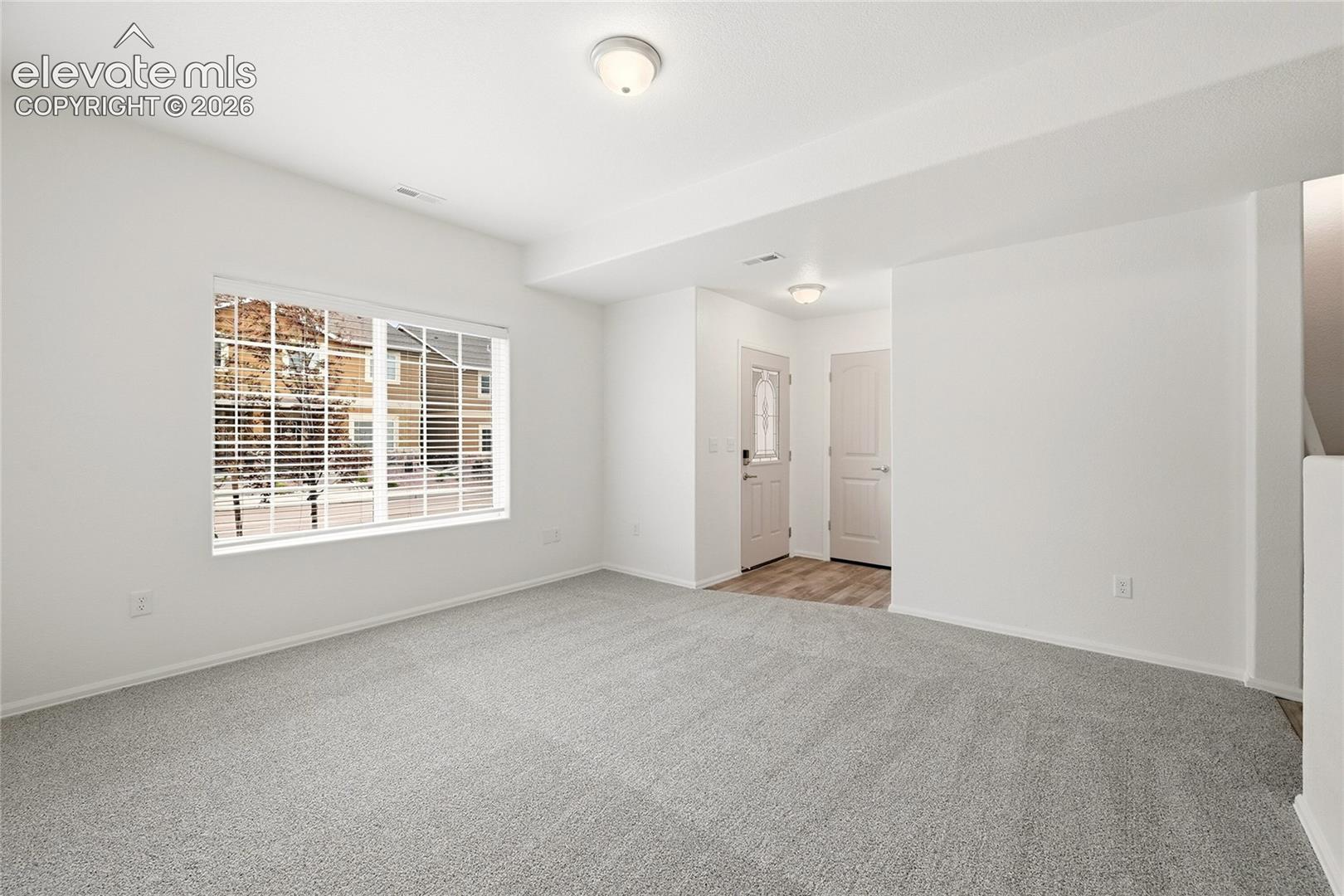 Neutral interior featuring wall-to-wall carpeting, white walls, flush-mount ceiling lighting, large picture window with blinds, and paneled doors