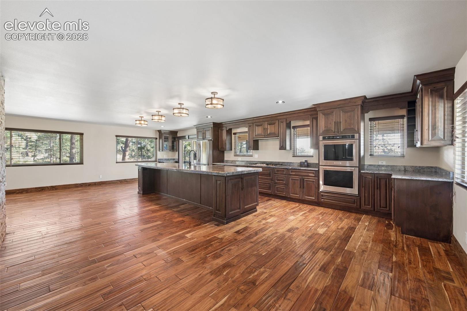 Kitchen featuring dark wood-finish cabinetry, stone countertops, stainless steel double wall ovens, wood-finish flooring, and multiple overhead flush-mount light fixtures