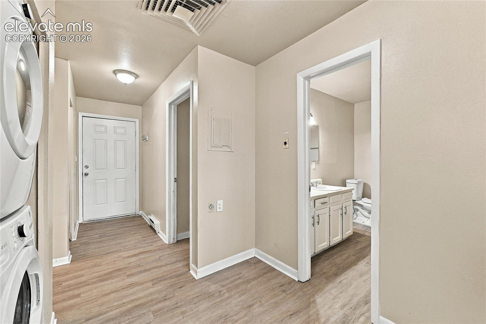 Utility area featuring wood-finish flooring, stackable appliance hookups, a six-panel door, and interior access to a bathroom with a white vanity