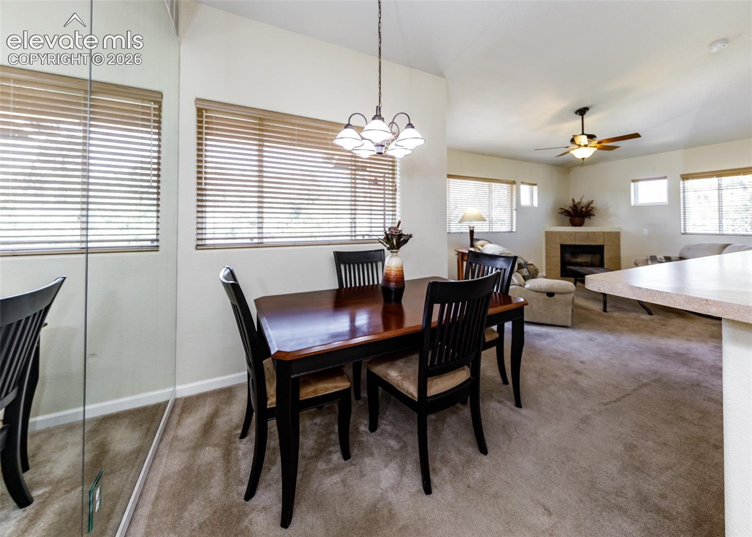 Dining area featuring a decorative chandelier, expansive mirrored wall, corner fireplace with tile surround, ceiling fan, and large windows with horizontal blinds