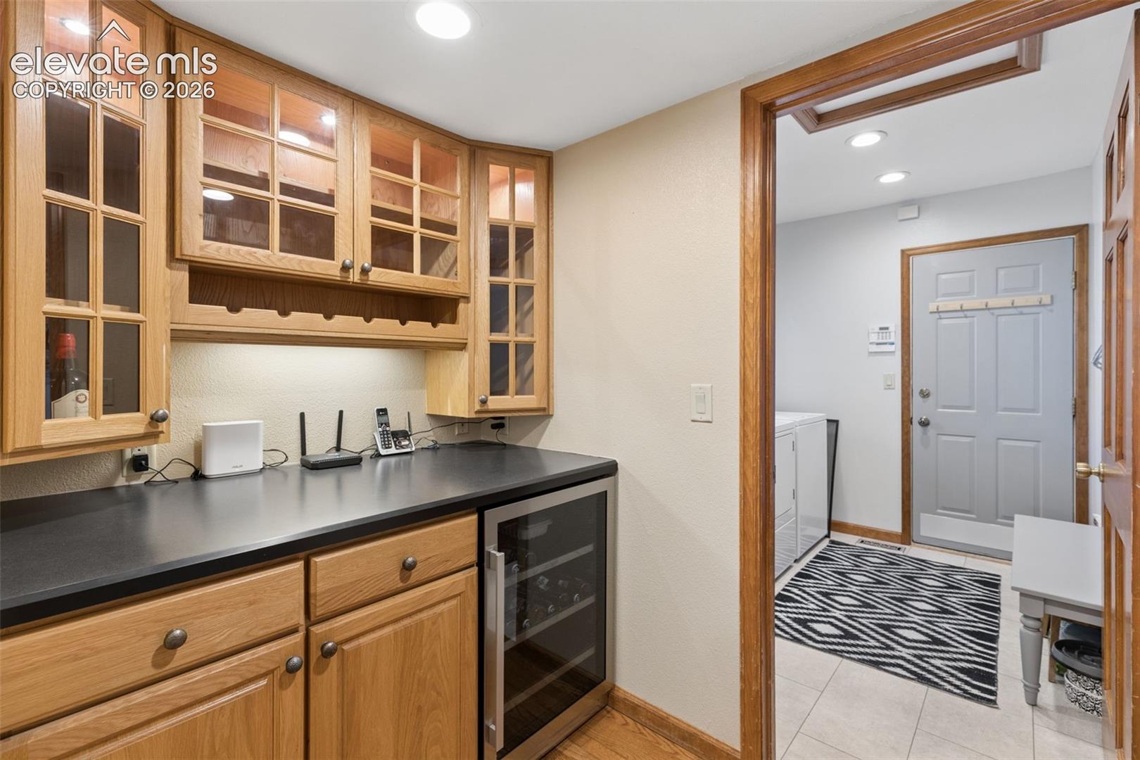 Custom cabinetry with glass-panel upper doors and integrated wine rack, dark solid-surface countertop, built-in stainless steel beverage refrigerator, and recessed lighting