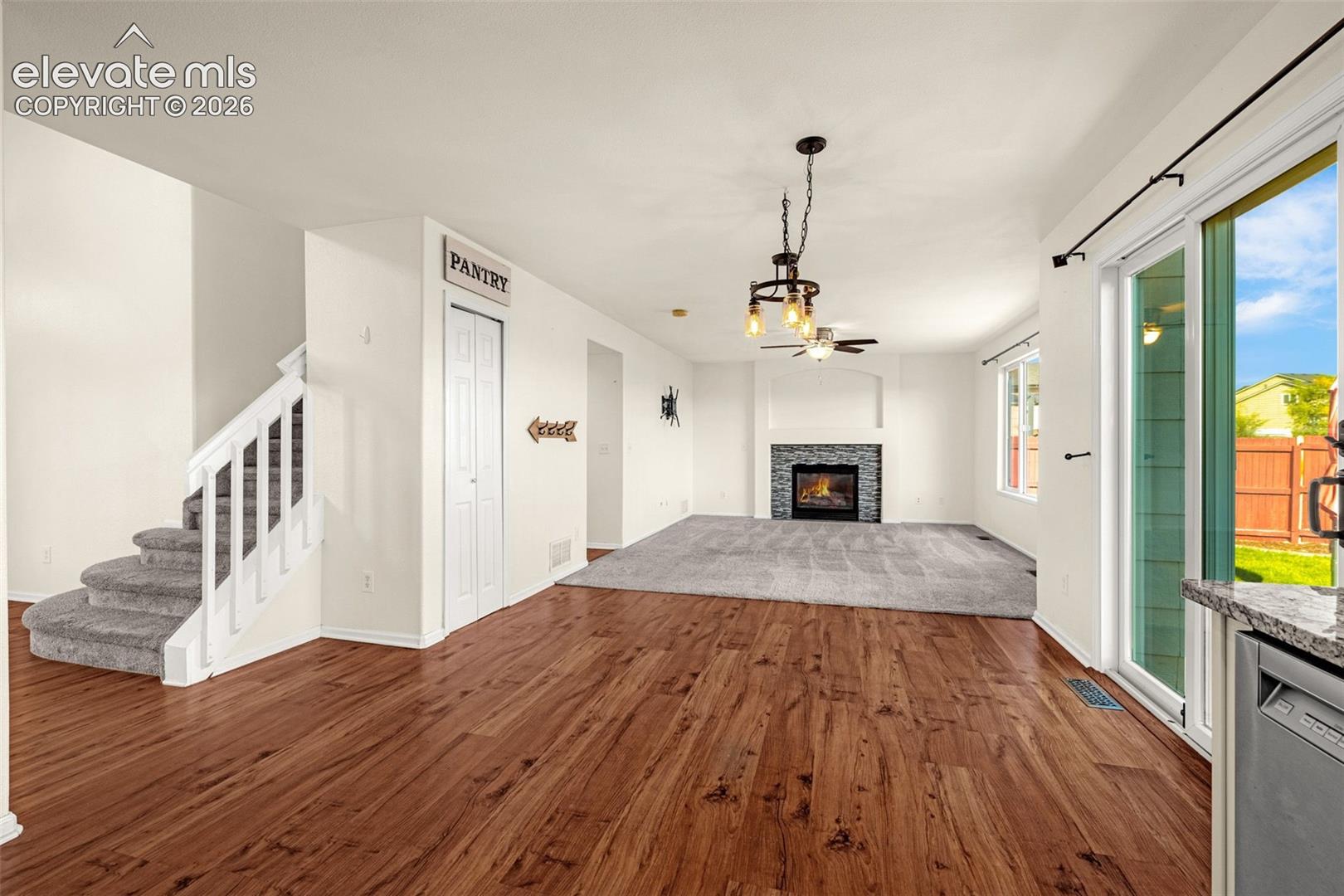 Open-concept interior featuring wood-finish flooring, a fireplace with a stone-finish surround, a multi-light metal chandelier, carpeted stairs with a white railing, and sliding glass doors