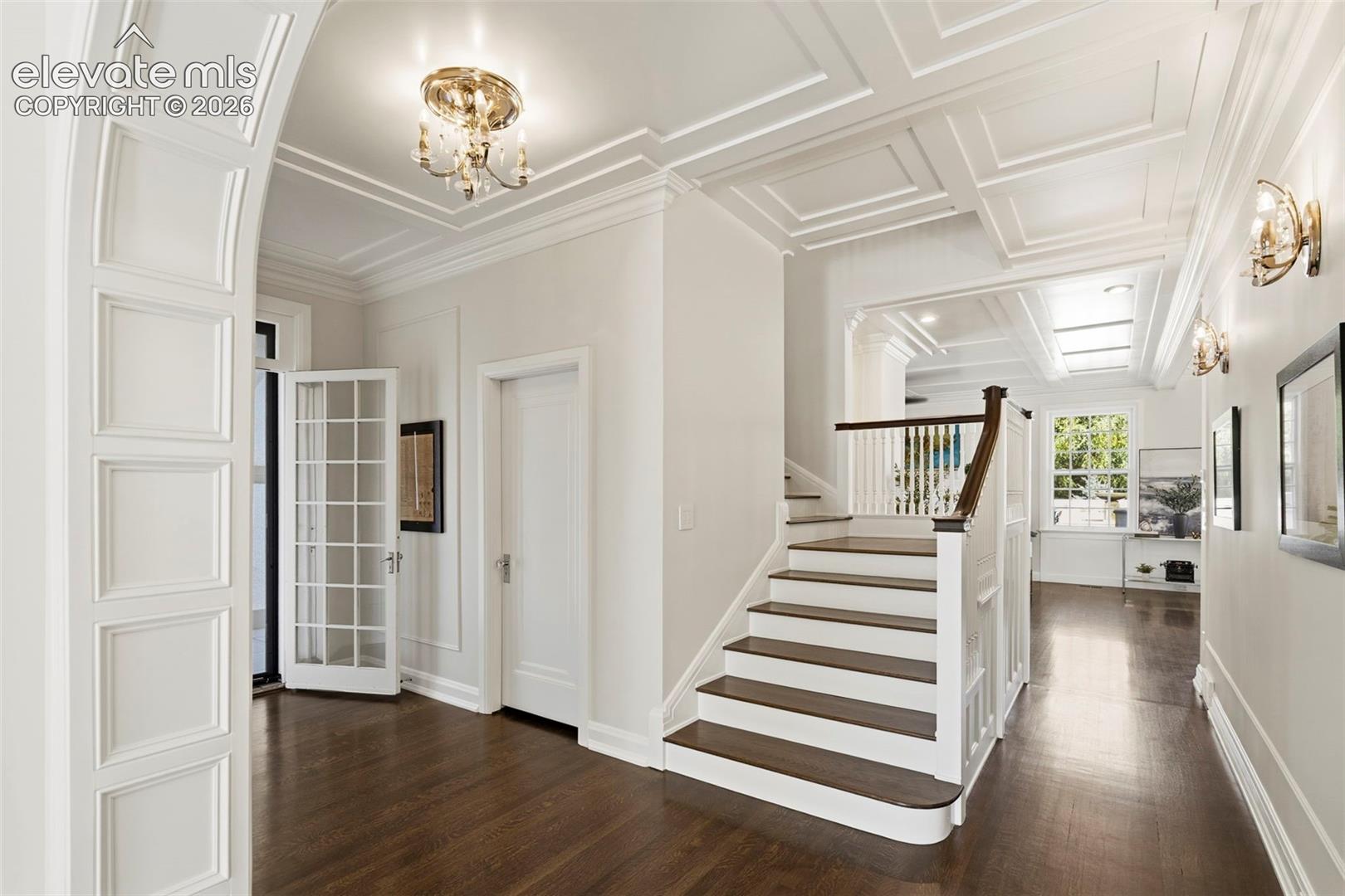 Foyer featuring a coffered ceiling, wood-finish flooring, an arched entryway with wainscoting detail, a staircase with a dark wood handrail, and integrated wall sconce lighting