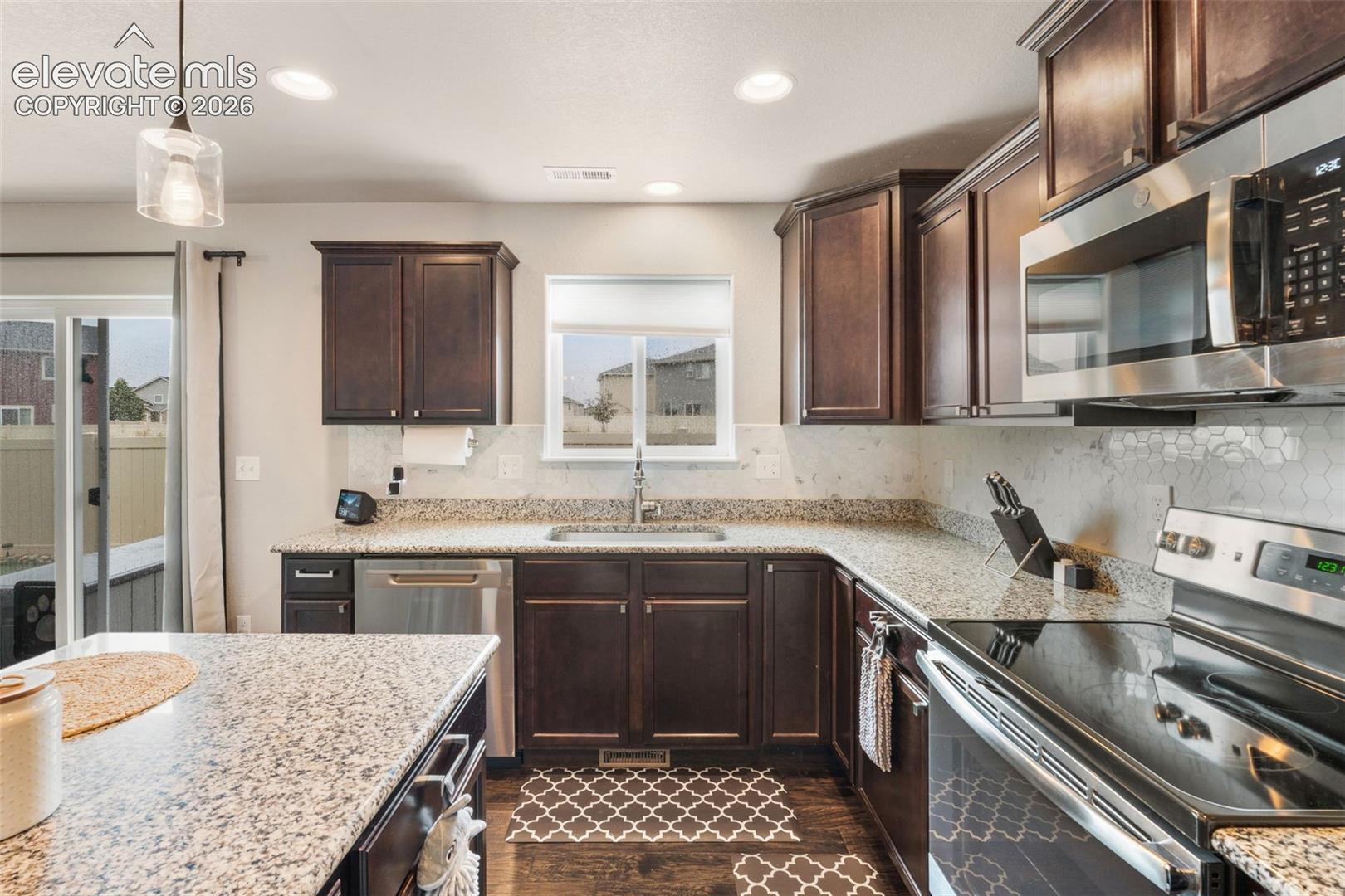 Kitchen featuring dark wood cabinetry, light granite countertops, stainless steel appliances, and a hexagon tile backsplash