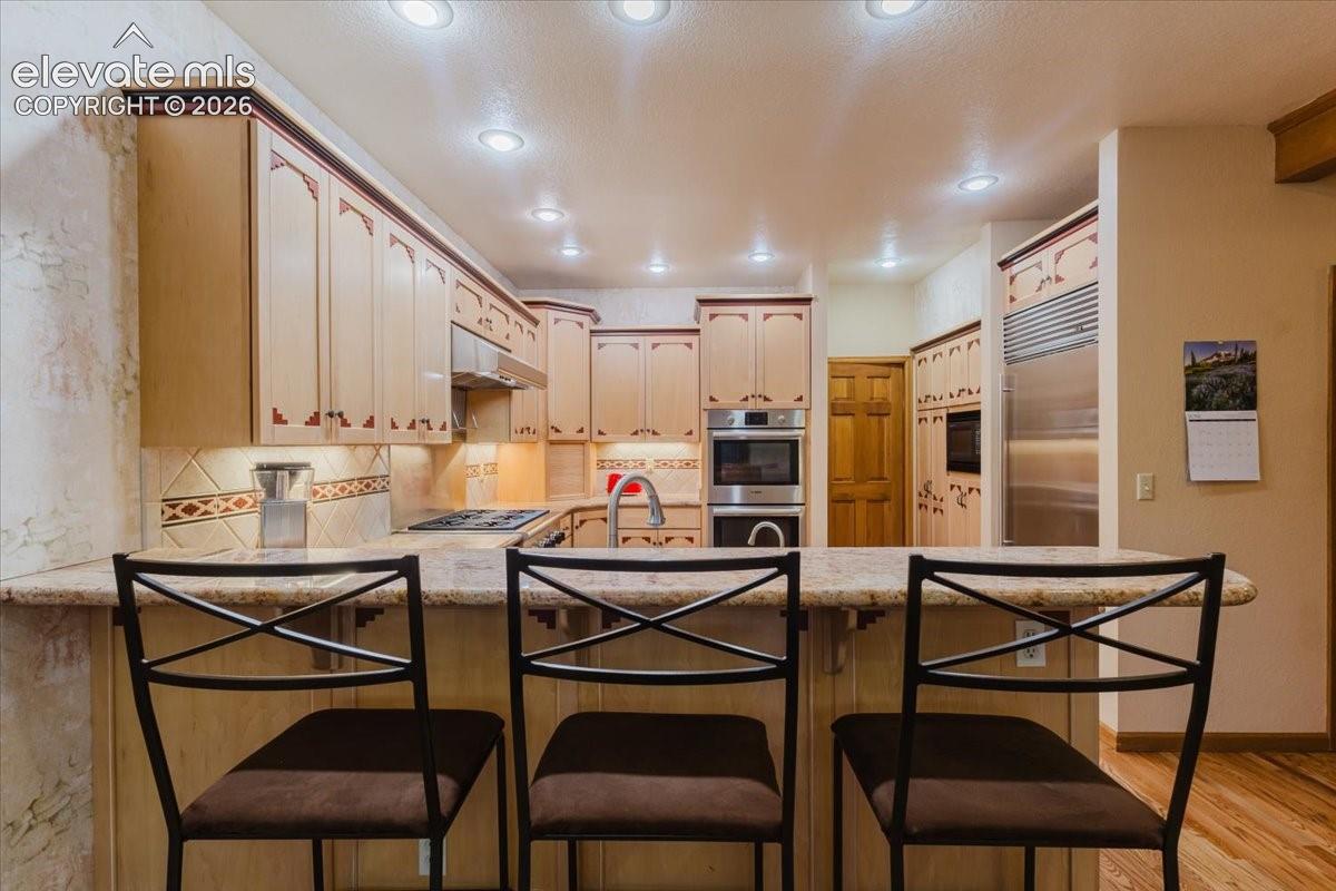 Kitchen featuring an expansive breakfast bar with stone-finish countertop, custom cabinetry, stainless steel appliances, recessed lighting, and a tiled backsplash