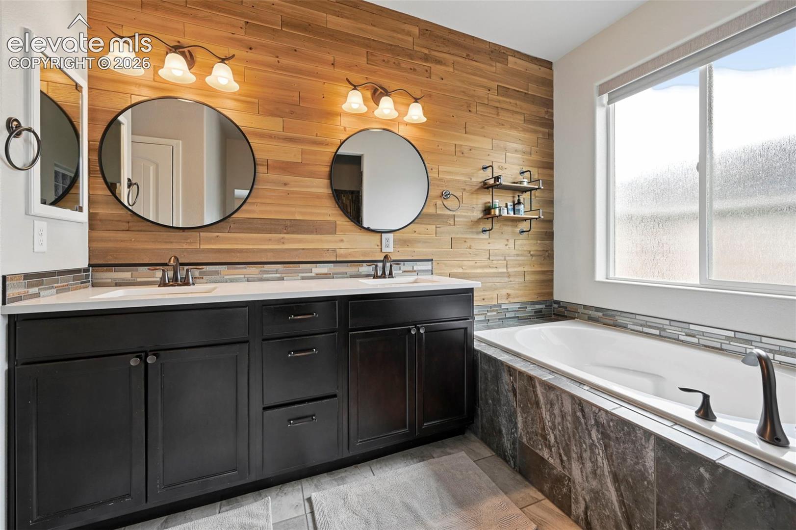 Double-vanity bathroom featuring a wood-plank accent wall, dark cabinetry, stone-tiled bathtub surround, and dual round mirrors with decorative wall-mounted lighting