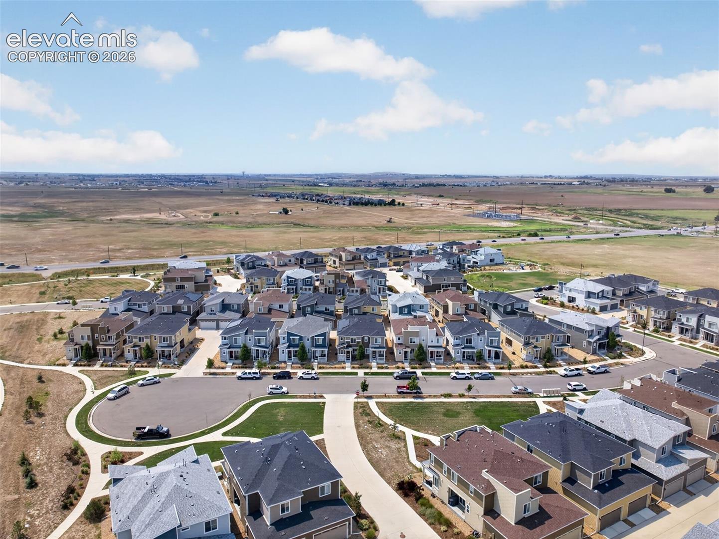 Aerial perspective of a suburban neighborhood featuring paved cul-de-sac streets, concrete sidewalks, multi-story residences with asphalt shingle roofs, and surrounding open plains