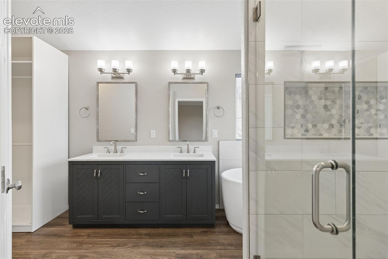 Double vanity with chevron-patterned cabinet doors, white countertop, dual undermount sinks, wood-finish flooring, and a walk-in shower with decorative mosaic tile inlay