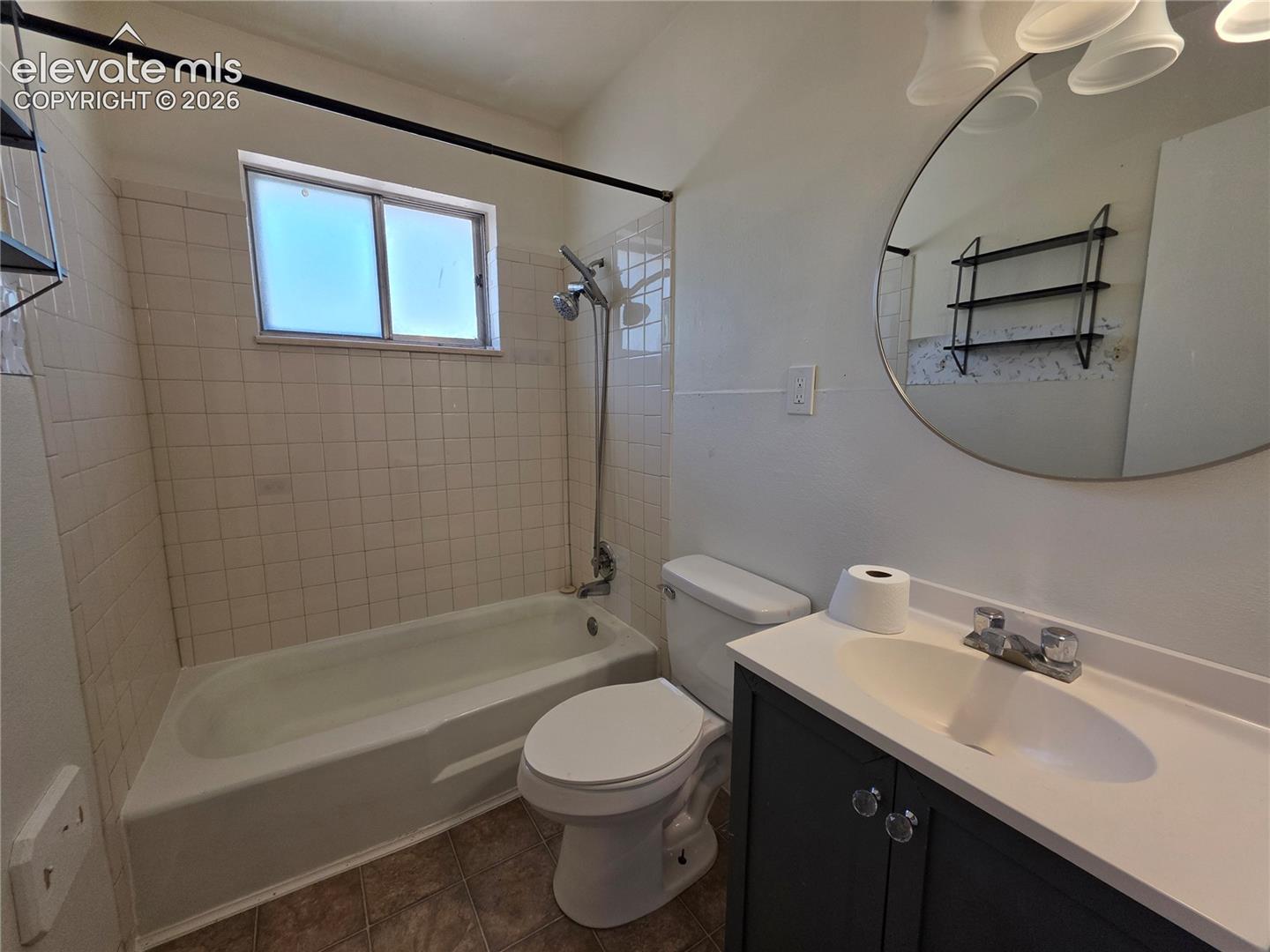 Full bathroom featuring a rectangular window, white tiled shower surround, a built-in tub, a dark gray vanity with a white countertop and integrated sink, and an overhead light fixture