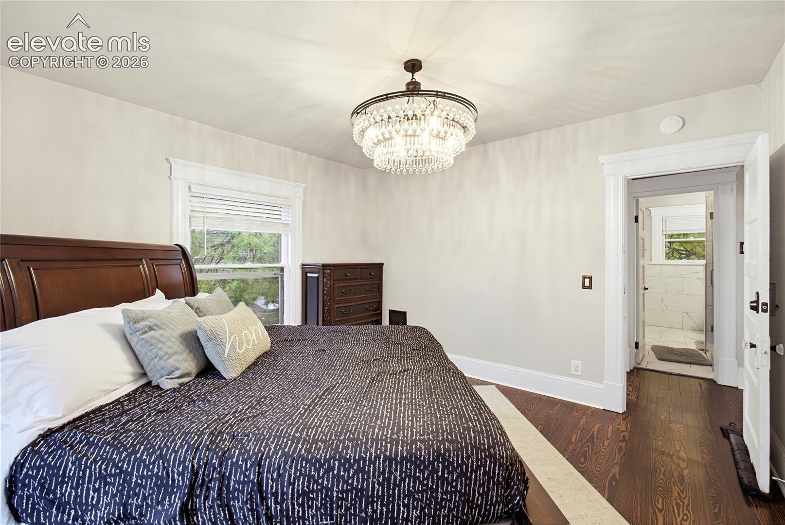 Hardwood flooring, elegant crystal chandelier, window with white blinds, and a doorway leading to a tiled bathroom